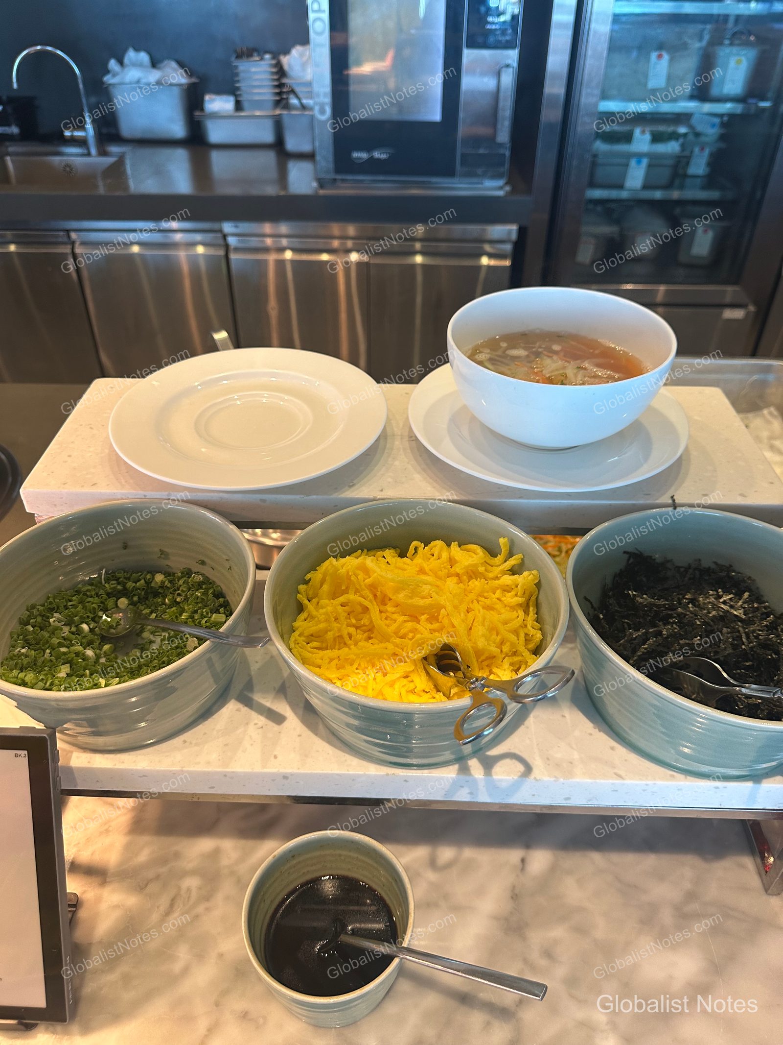 Noodle soup station at the Grand Hyatt Jeju breakfast buffet, featuring neatly arranged bowls of garnishes like chopped green onions, shredded egg strips, and dried seaweed, alongside bowls of hot broth and empty serving plates.