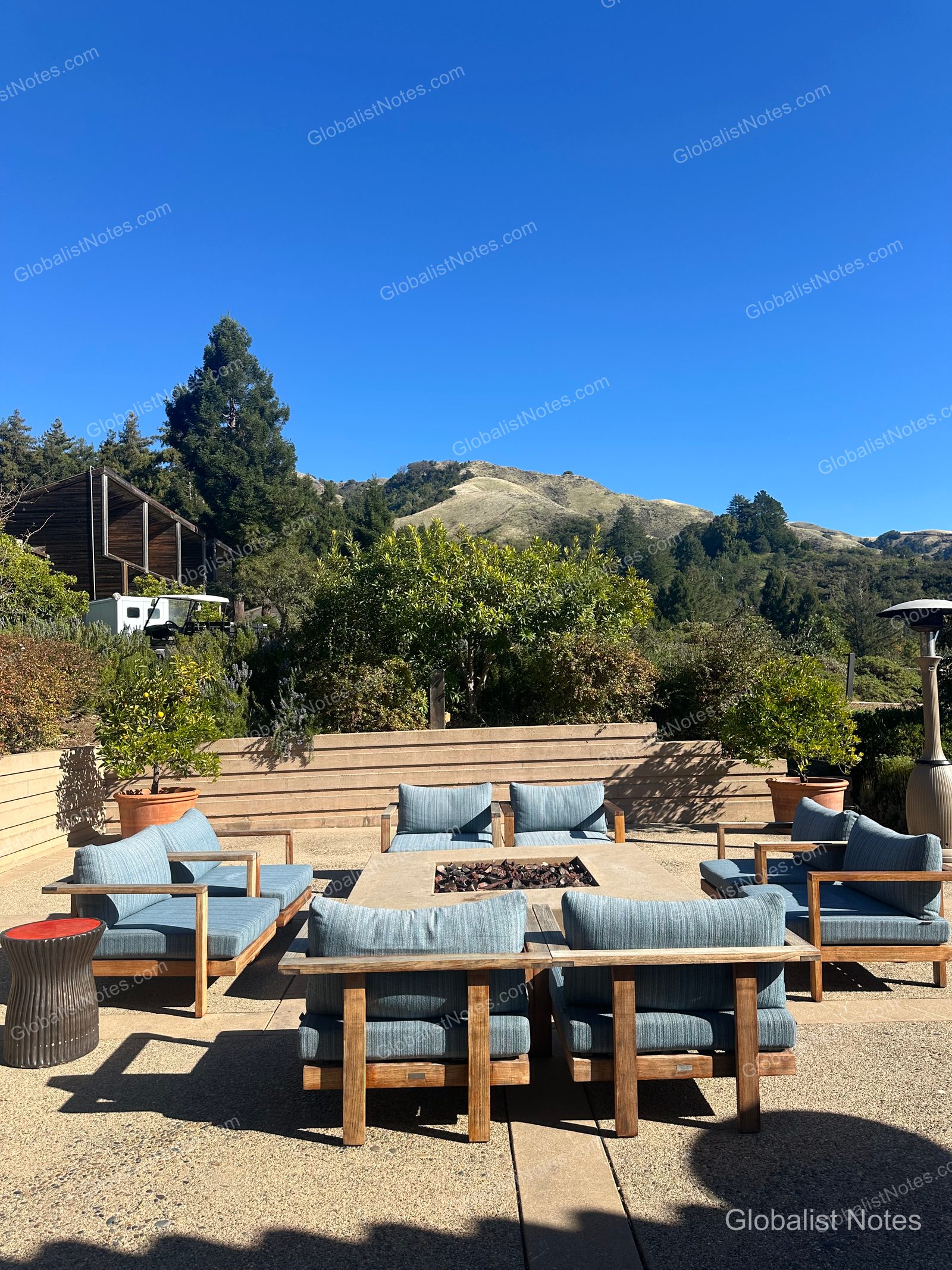 Wooden chairs with blue cushions arranged around a fire pit on the outdoor terrace at Alila Ventana Big Sur, with green mountains and trees visible in the background under a clear sky.