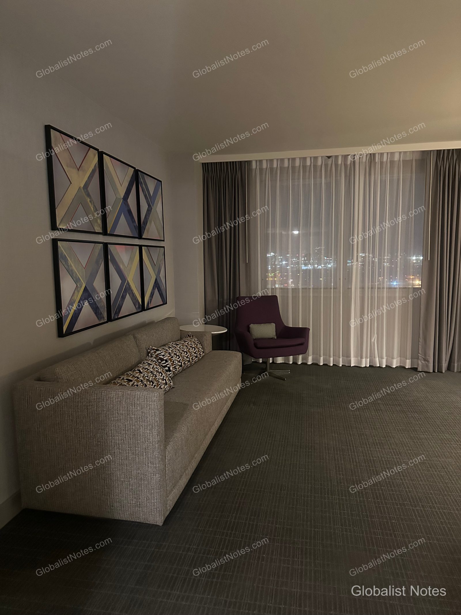 Living room of a One Bedroom King Suite at Hyatt Regency Los Angeles International Airport. The space features a gray sofa, a purple armchair, and four geometric art pieces on the wall, with a view of the Los Angeles night skyline through the window.