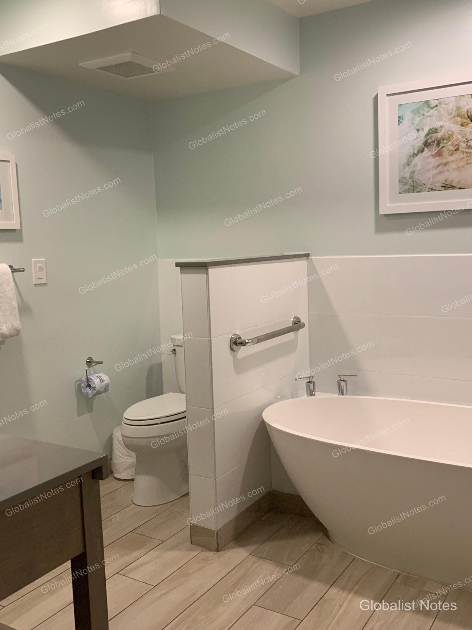 Hyatt Vacation Club at Sunset Harborの客室バスルーム。淡いミントグリーンの壁に明るい木目調のタイル床が映え、白いバスタブとトイレが低いパーティションで仕切られています。壁には海の風景画が飾られ、バスタブには手すりが備わっています。