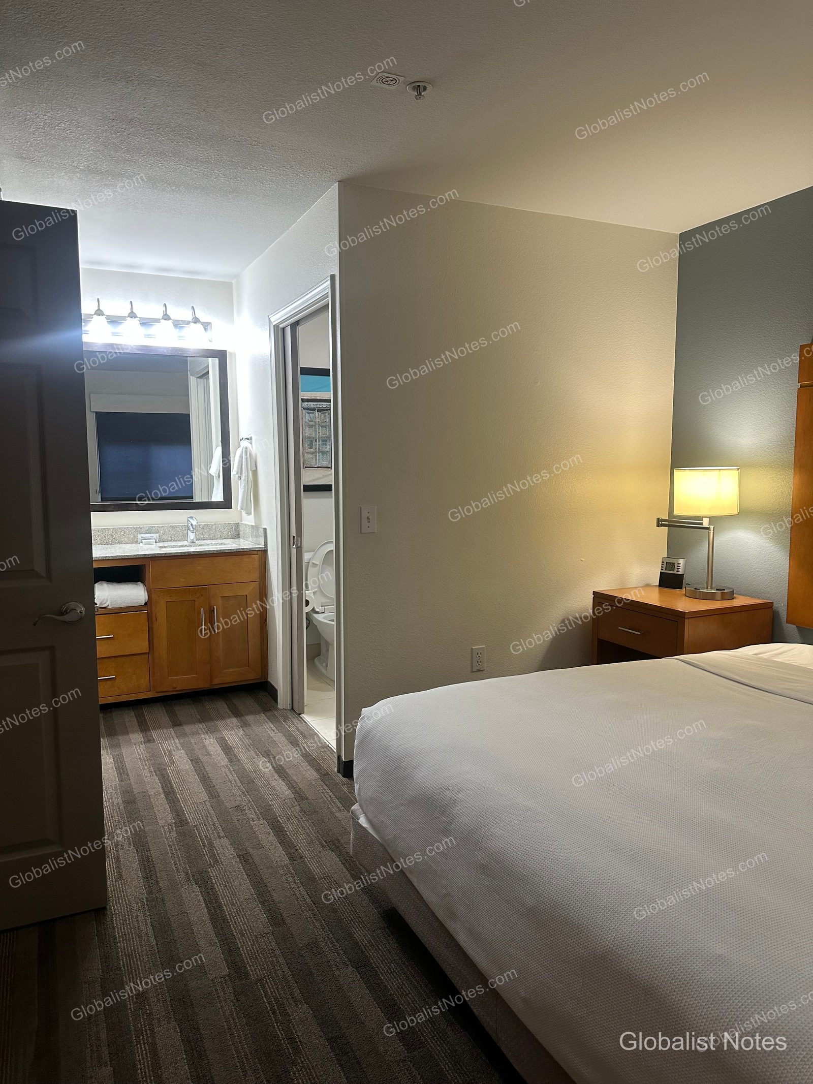 Hyatt House Scottsdale/Old Townのワンベッドルームスイートの室内。ベッドサイドのナイトテーブルと照明、その奥に洗面エリアが見える。
