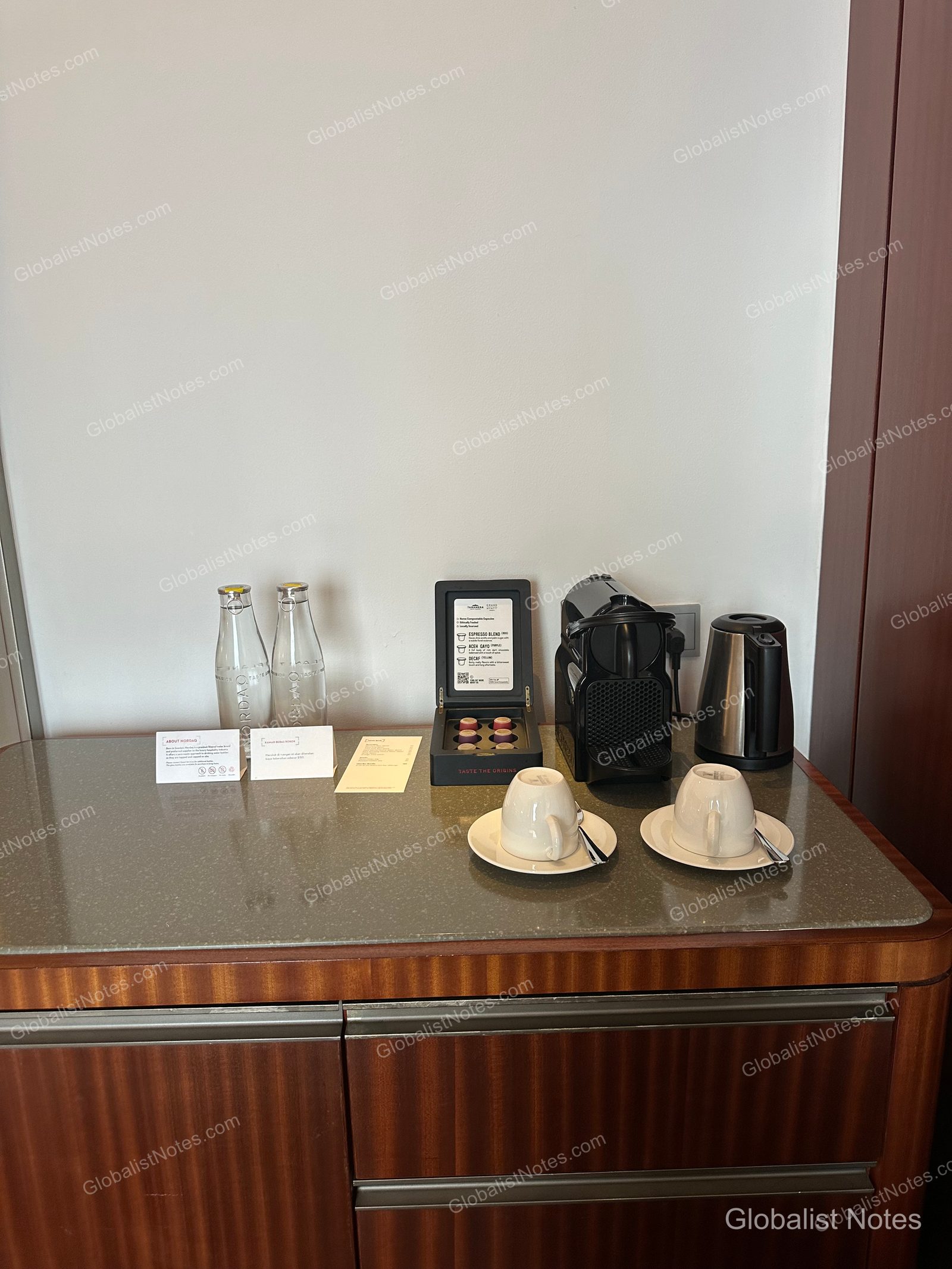 Minibar area in a Grand Hyatt Jakarta guest room, featuring a capsule coffee machine, electric kettle, two glass bottles of water, and a set of coffee cups arranged on a marble countertop.