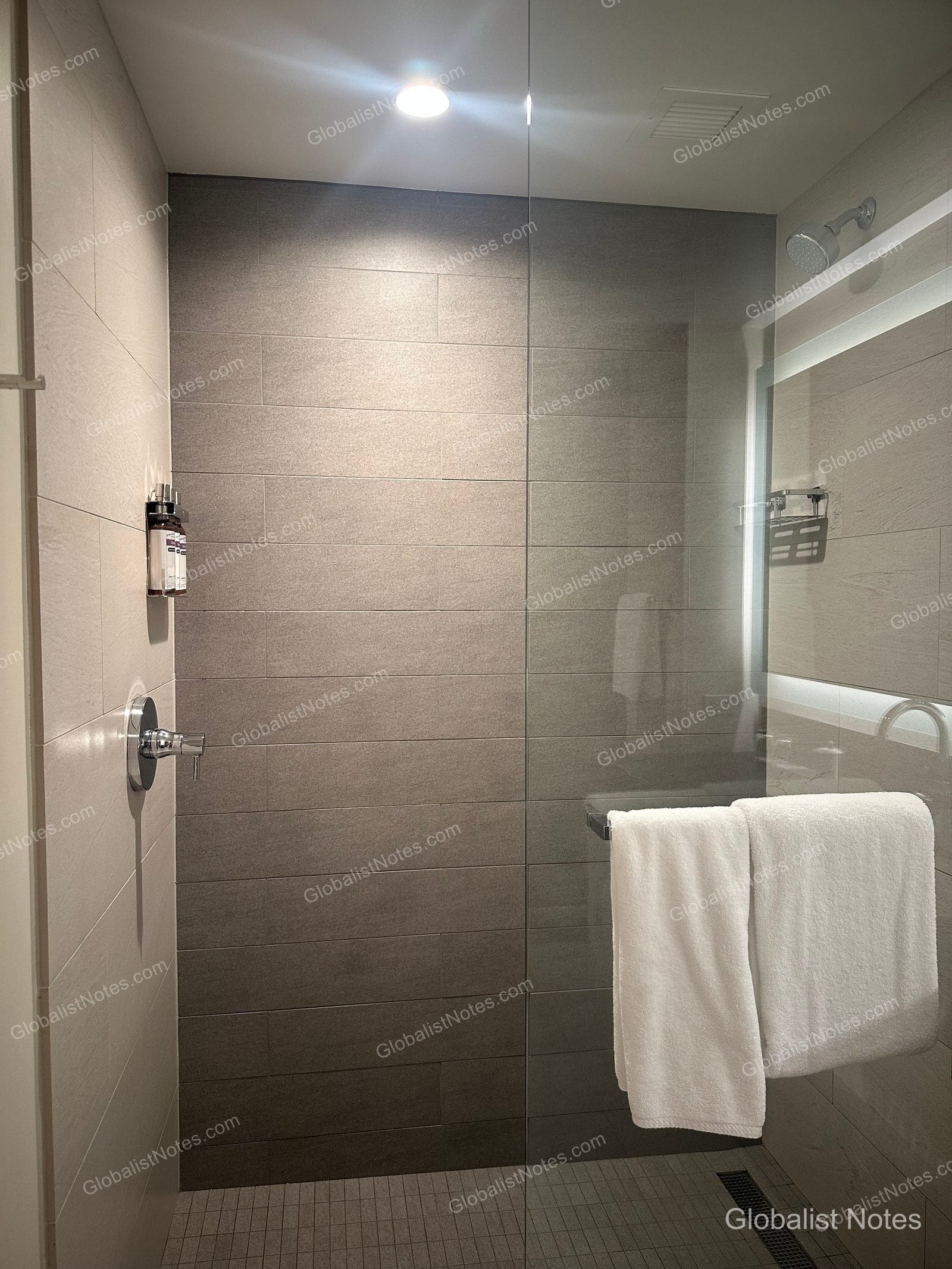Bathroom interior of a standard king room at Hyatt Regency Seattle. The shower stall is finished with gray tiles and features a glass partition, wall-mounted amenity dispensers, and a towel rack, all illuminated by ceiling lighting.
