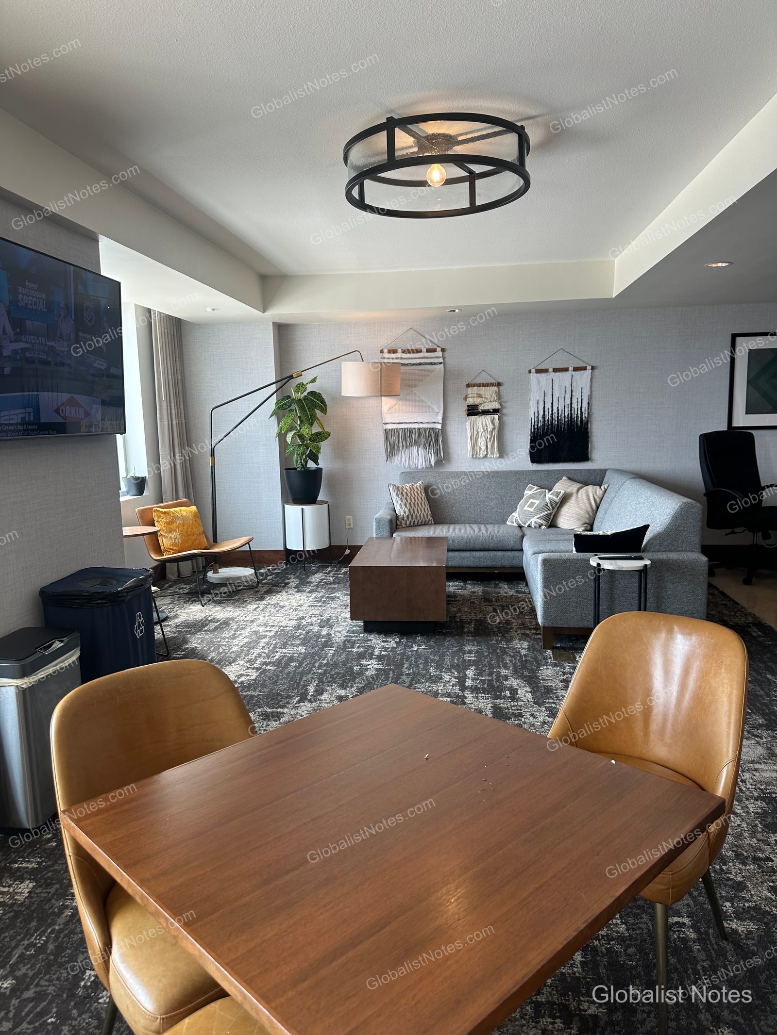 The living room of an Executive Suite at Grand Hyatt San Antonio River Walk. It features a gray L-shaped sofa, a wooden coffee table, a decorative wall tapestry, a wooden dining table with leather chairs, and a wall-mounted TV.