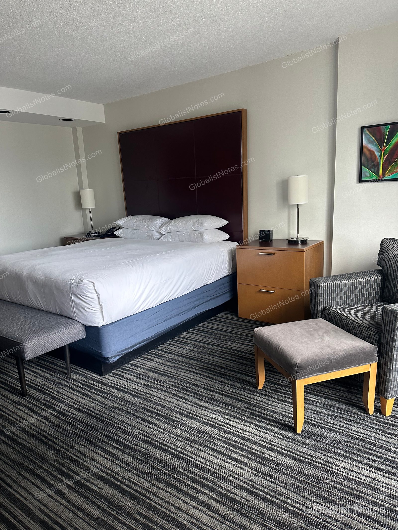 Bedroom view of the Deluxe Suite at Hyatt Regency Vancouver. A king bed with a dark brown headboard is flanked by nightstands and lamps, with a gray bench at the foot of the bed and a plaid armchair with an ottoman set on a striped carpet to the right.