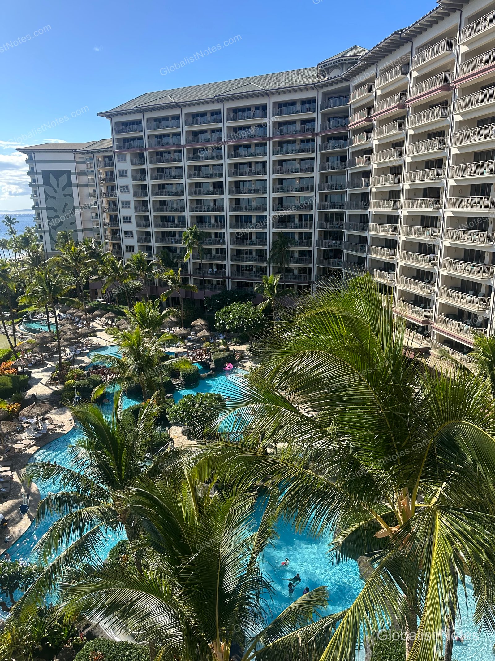 View of Hyatt Regency Maui Resort and Spa in Lahaina, Hawaii. The resort features a pool area with palm trees and umbrellas in front of the main building, with guests swimming in the pool.
