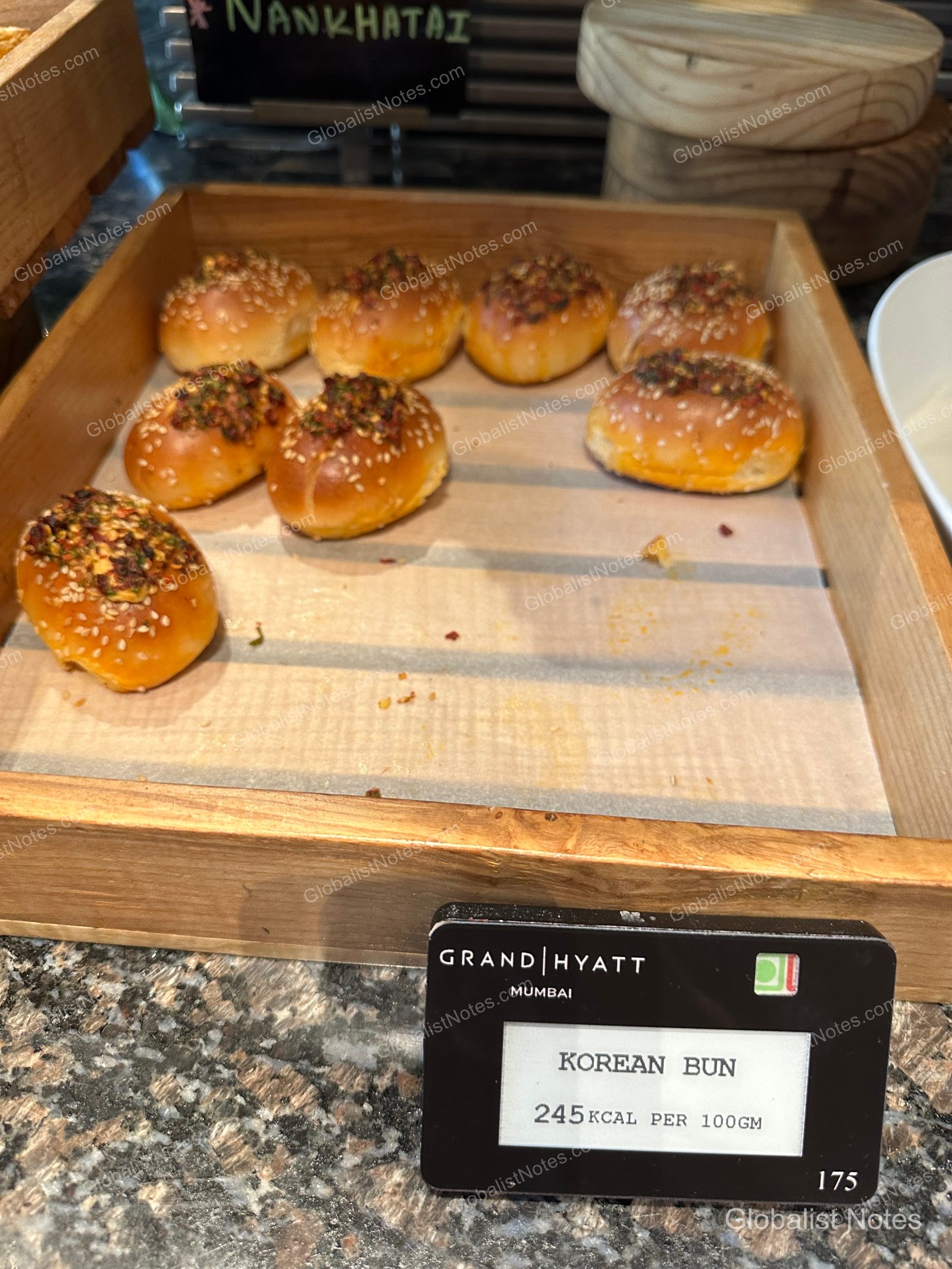 A wooden tray at the Grand Hyatt Mumbai breakfast buffet holding golden-brown Korean-style buns topped with sesame seeds, with a small black sign labeled 'KOREAN BUN' in front.