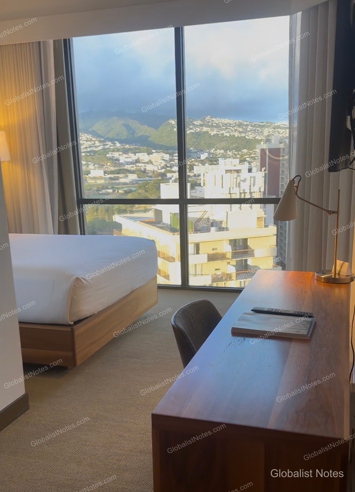 Interior of a guest room at Hyatt Centric Waikiki Beach, featuring floor-to-ceiling windows with views of downtown Honolulu and the mountain range. The room includes a bed with white linens, a wooden desk, and a floor lamp, with bright natural light streaming in.