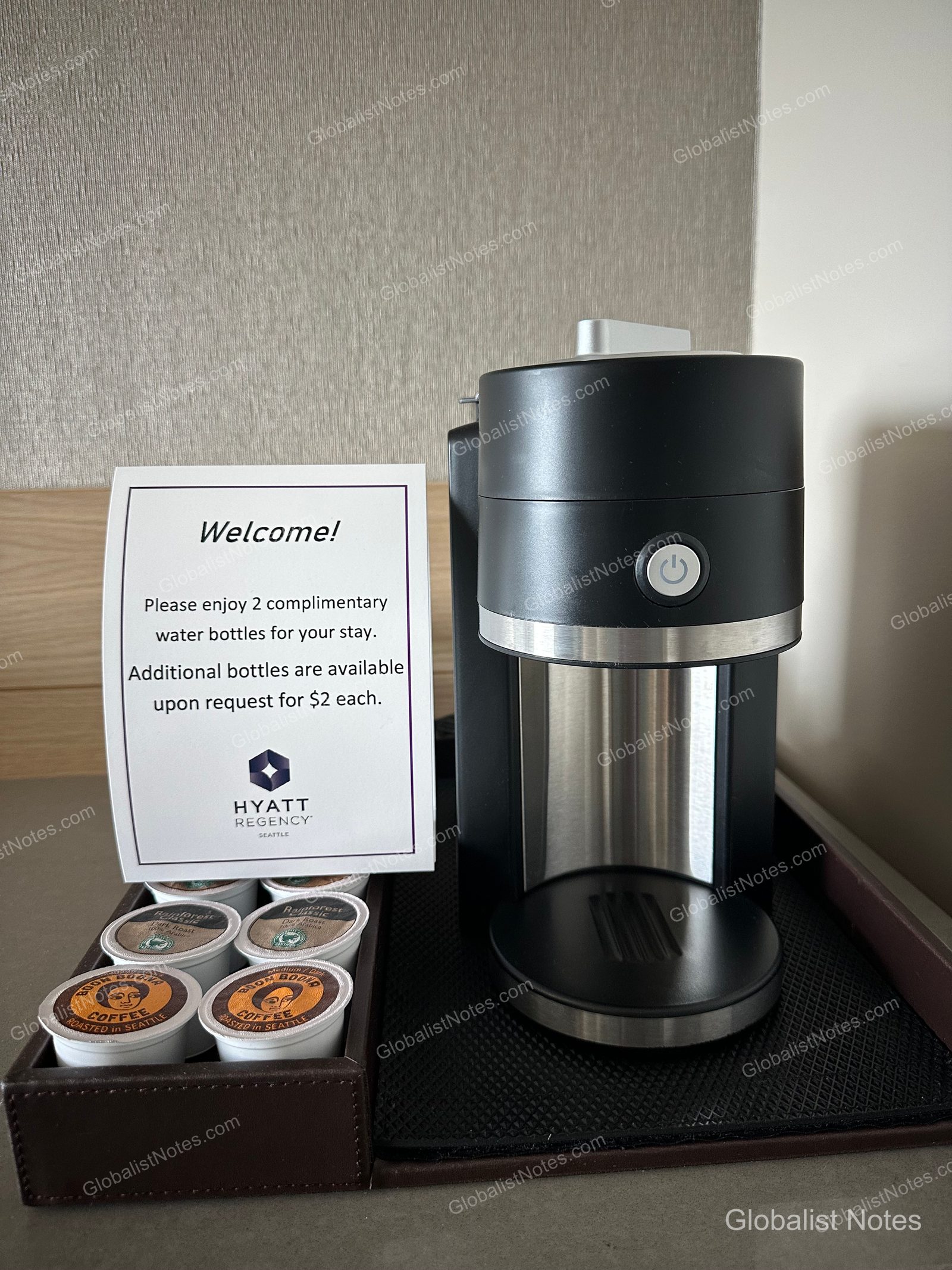 A black coffee machine and a tray of capsules are set up in a Hyatt Regency Seattle guest room, alongside a notice regarding bottled water. The notice states that two bottles of water are provided complimentary, with a $2 charge per additional bottle.