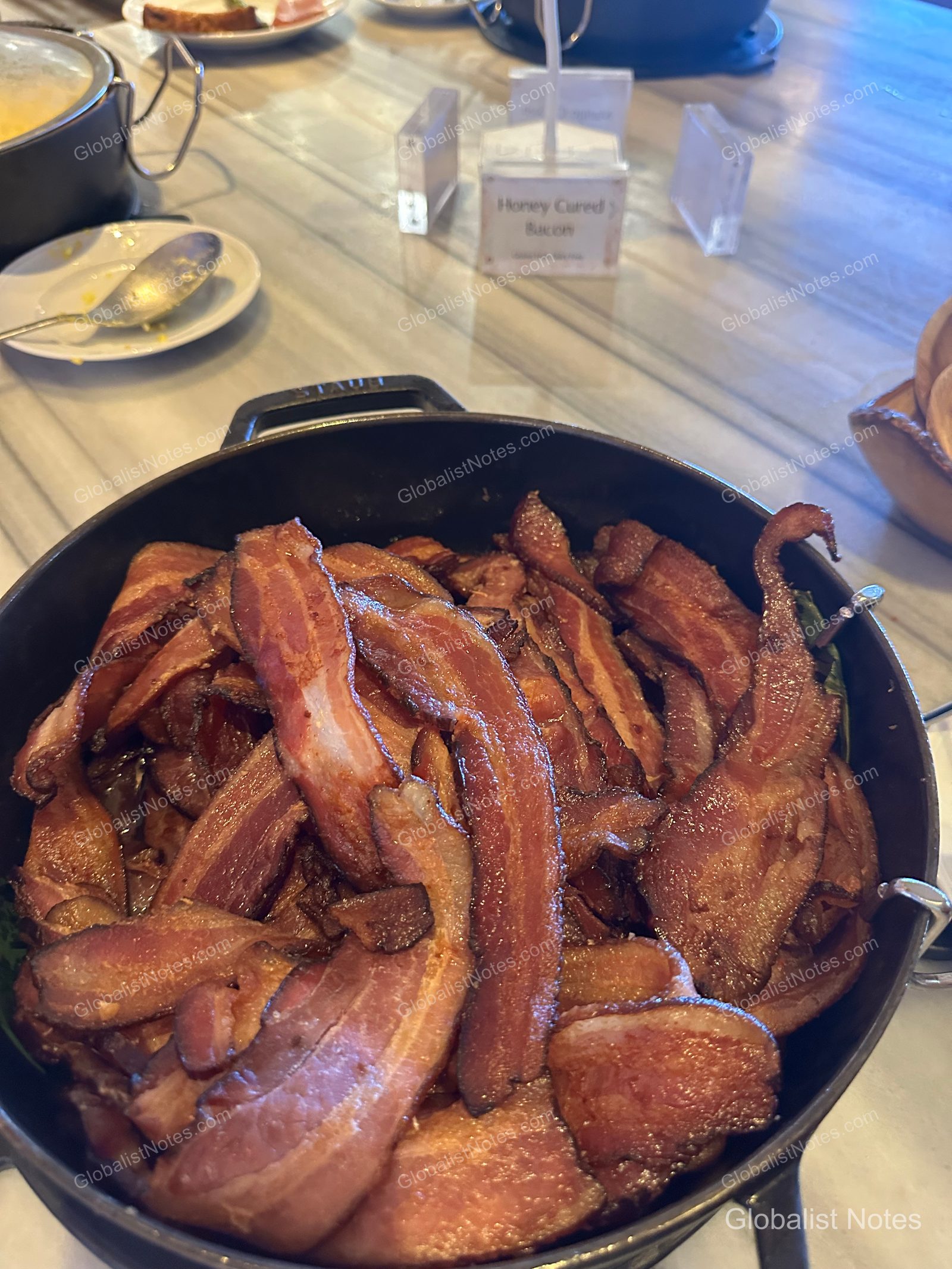 안다즈 마우이 앳 와일레아 리조트 조식 뷔페에 놓인 스타우브 무쇠 냄비에 허니 큐어드 베이컨이 가득 담겨 있습니다. 뒤쪽에는 메뉴 이름표가 보이며, 대리석 테이블 위에 정갈하게 세팅된 모습입니다.