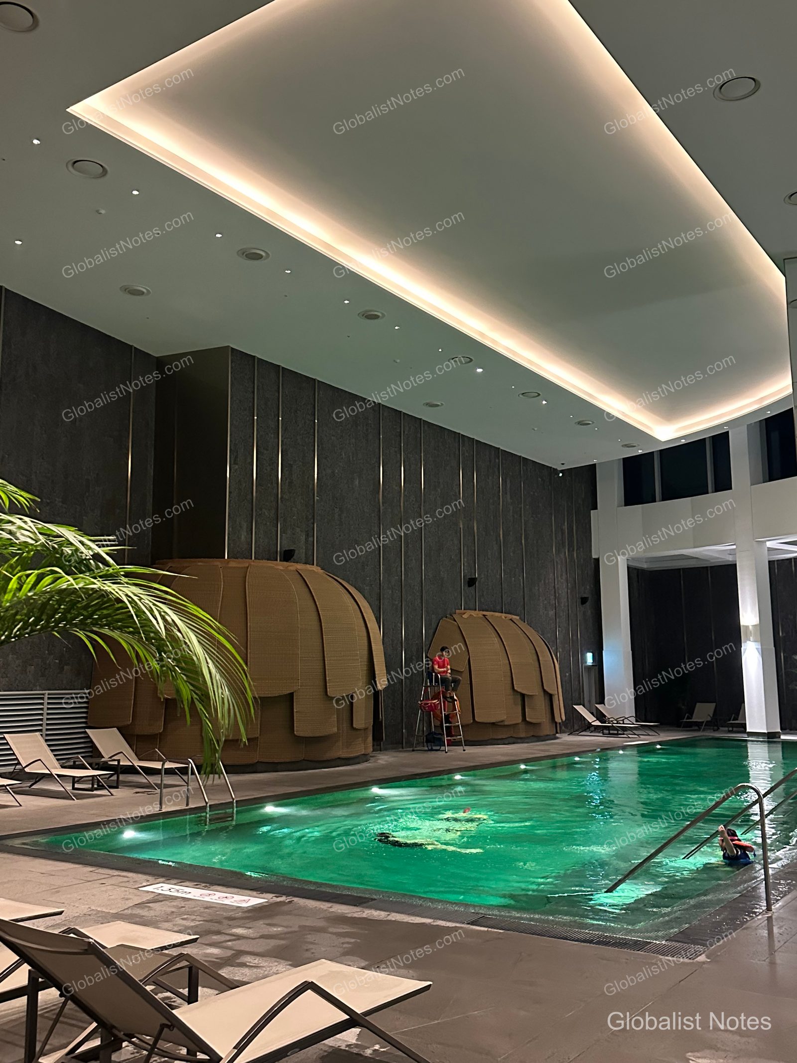 View of the indoor pool at Grand Hyatt Jeju, featuring a high ceiling with indirect lighting, a green-toned pool, dome-shaped cabanas, and neatly arranged sun loungers. Potted plants are placed around the pool area, with lifeguards on duty and guests swimming.