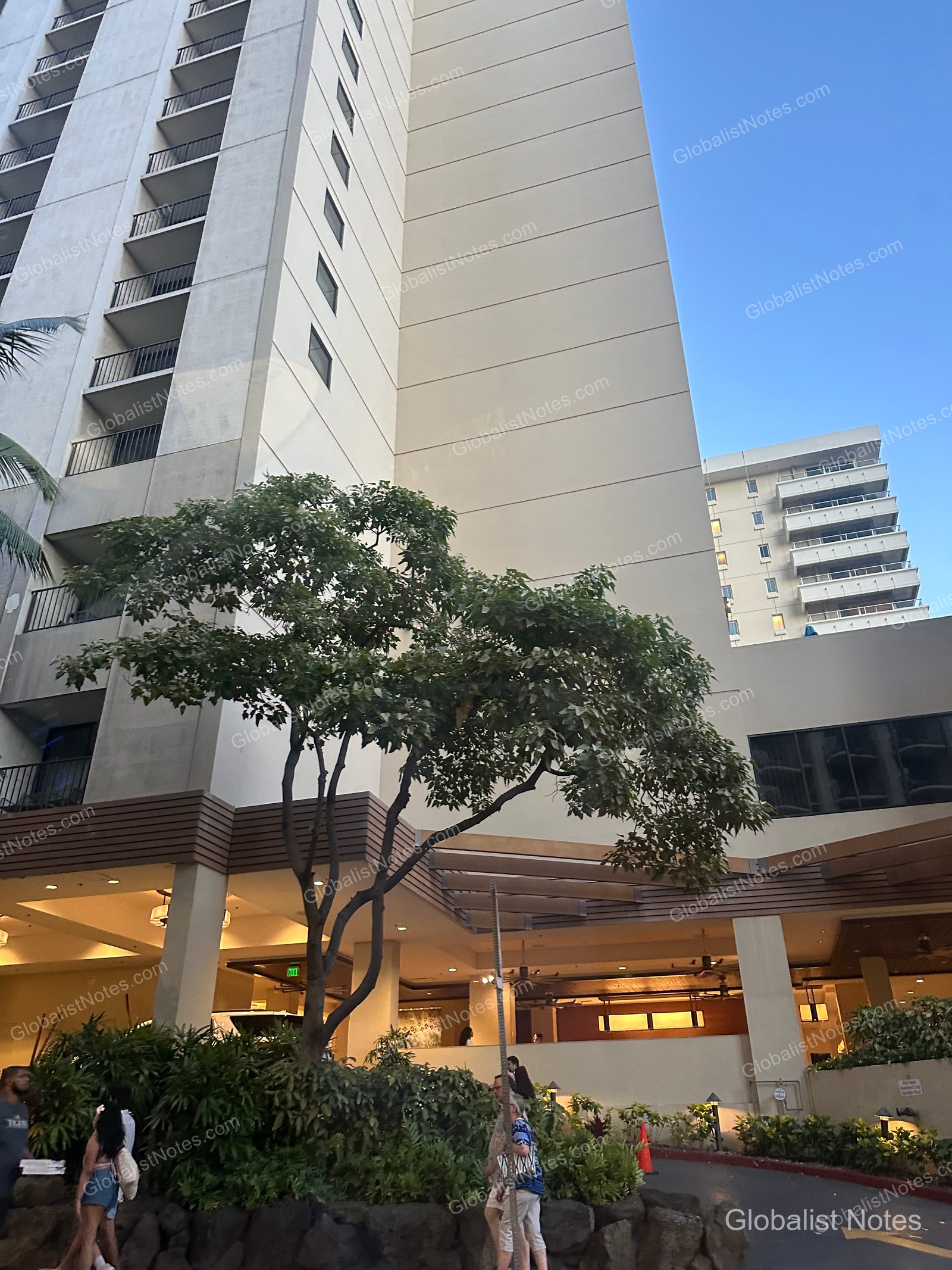 Exterior view of the Hyatt Place Waikiki Beach hotel in Honolulu, Hawaii, showing the building facade, landscaping near the entrance, and guests walking toward the lobby.