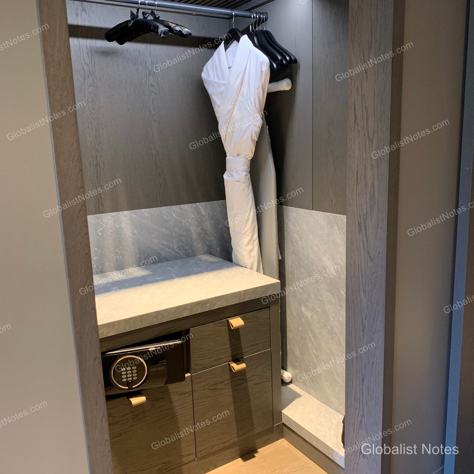Open-concept closet in an Alila Napa Valley guest room. White bathrobes hang from the top rack, while the lower section features a marble-topped cabinet with a built-in electronic safe. The space uses a neutral palette of wood and marble.