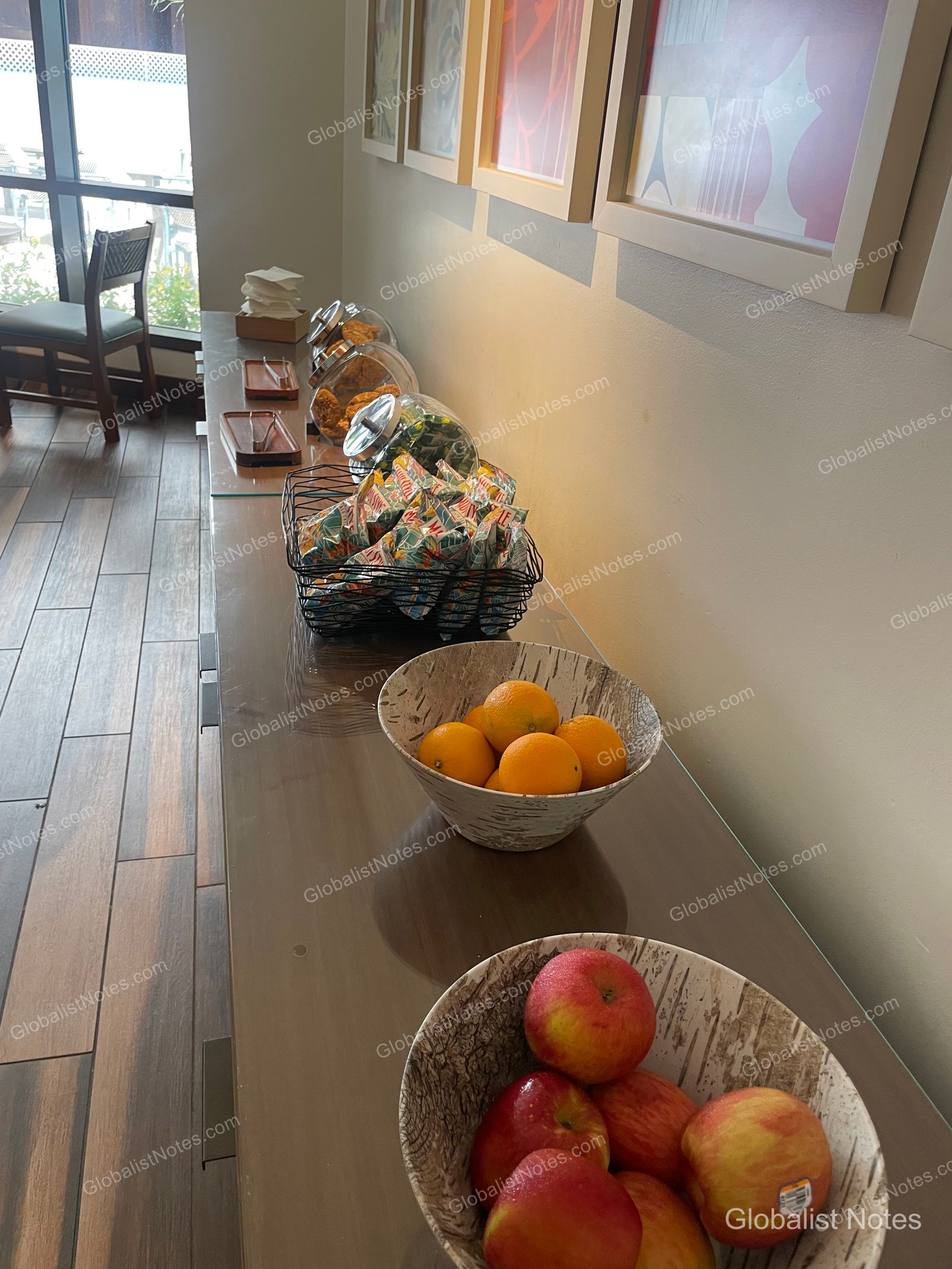 Snack station on a long table inside the lounge at Hyatt Regency Maui Resort and Spa, featuring individually wrapped snacks in baskets, cookies in glass jars, and oranges and apples in wooden bowls.