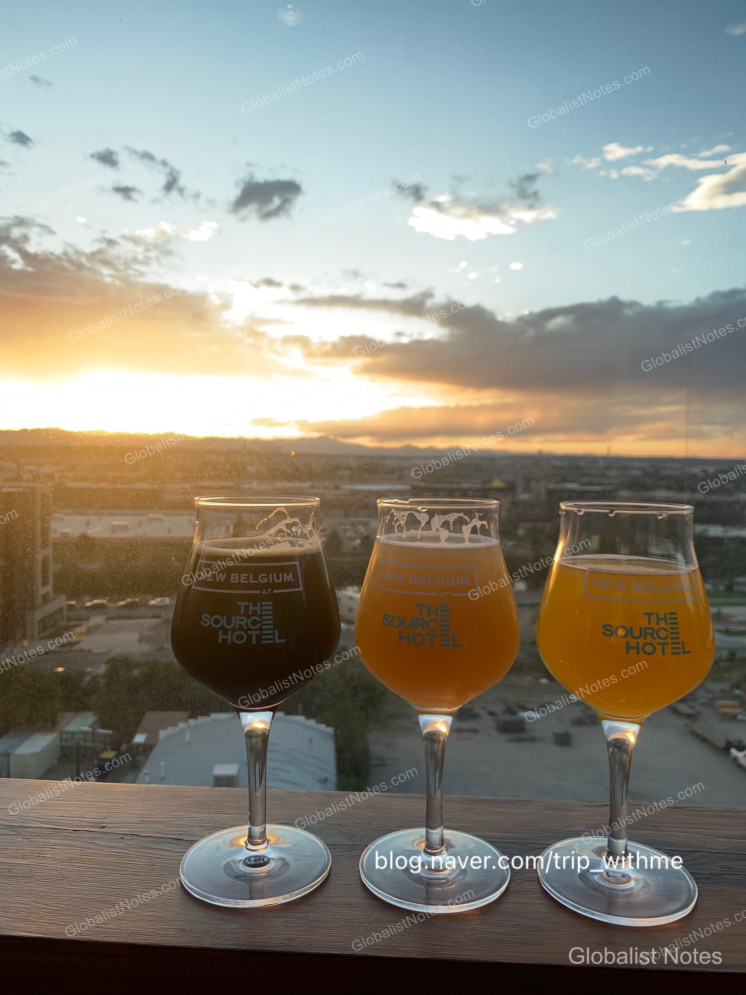 デンバーの夕暮れを背景に、窓際のテーブルに並べられた「The Source Hotel」のロゴ入りビールグラス。それぞれ異なる色のビールが注がれ、窓の外には山々と街の全景が広がっている。