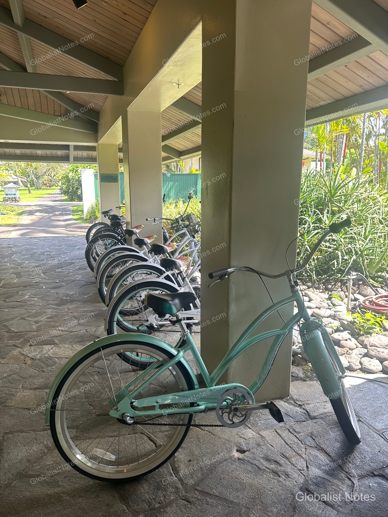 Mint-colored bicycles lined up along the outdoor corridor pillars at Hana-Maui Resort, with the resort's lush gardens and walking paths visible in the bright sunlight beyond.
