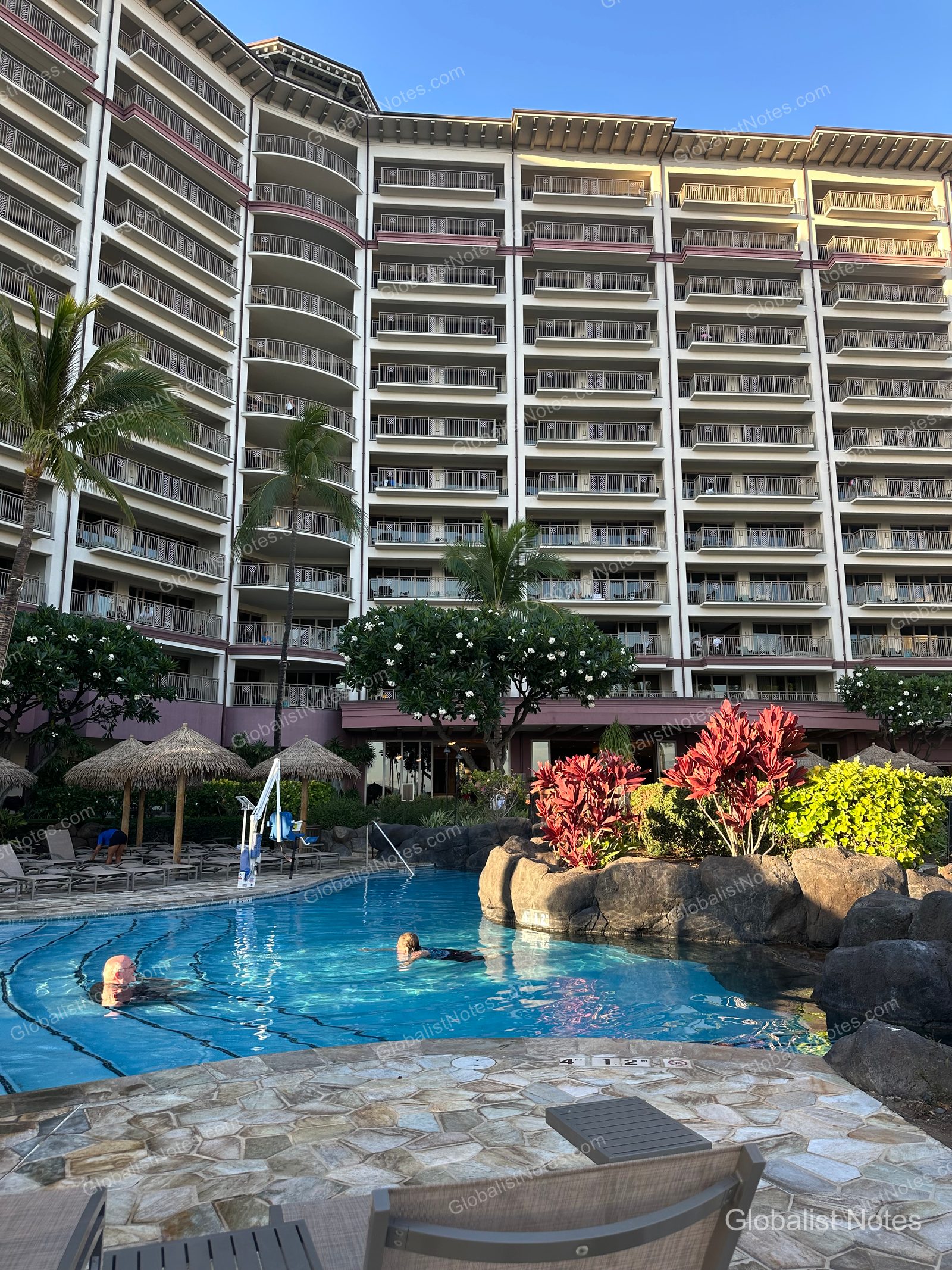 Exterior view of Hyatt Regency Maui Resort and Spa in Lahaina, Hawaii. The multi-story guest room buildings overlook an outdoor pool area landscaped with palm trees and rock features, with umbrellas and sun loungers arranged around the pool.