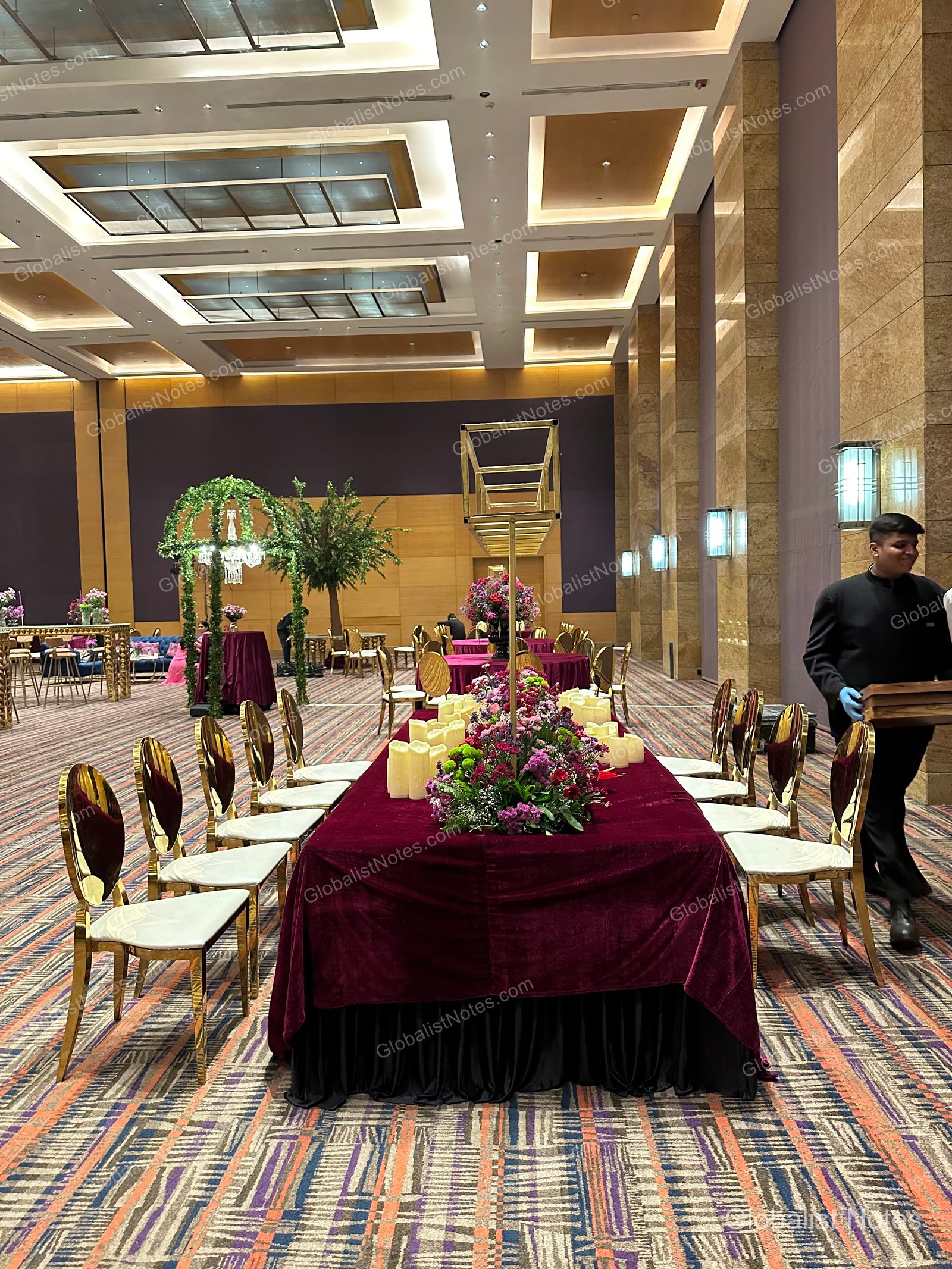 A spacious banquet hall at Grand Hyatt Mumbai with long tables draped in deep purple linens, accented by floral arrangements and gold chairs. A staff member in a black uniform walks across the frame beneath a grid-patterned ceiling light design.