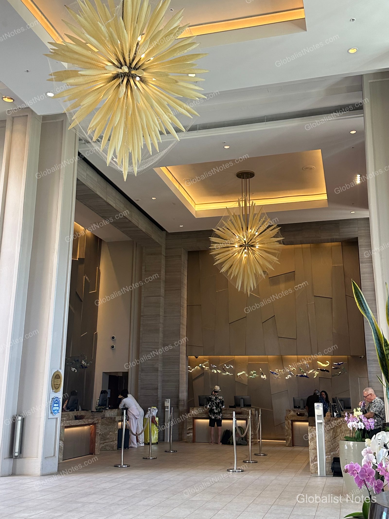View of the open-concept lobby at Hyatt Regency Maui Resort and Spa in Lahaina, Hawaii, featuring high ceilings and large sculptural light fixtures. Guests are standing at the check-in counters, and the floor is finished with light-toned tiles.