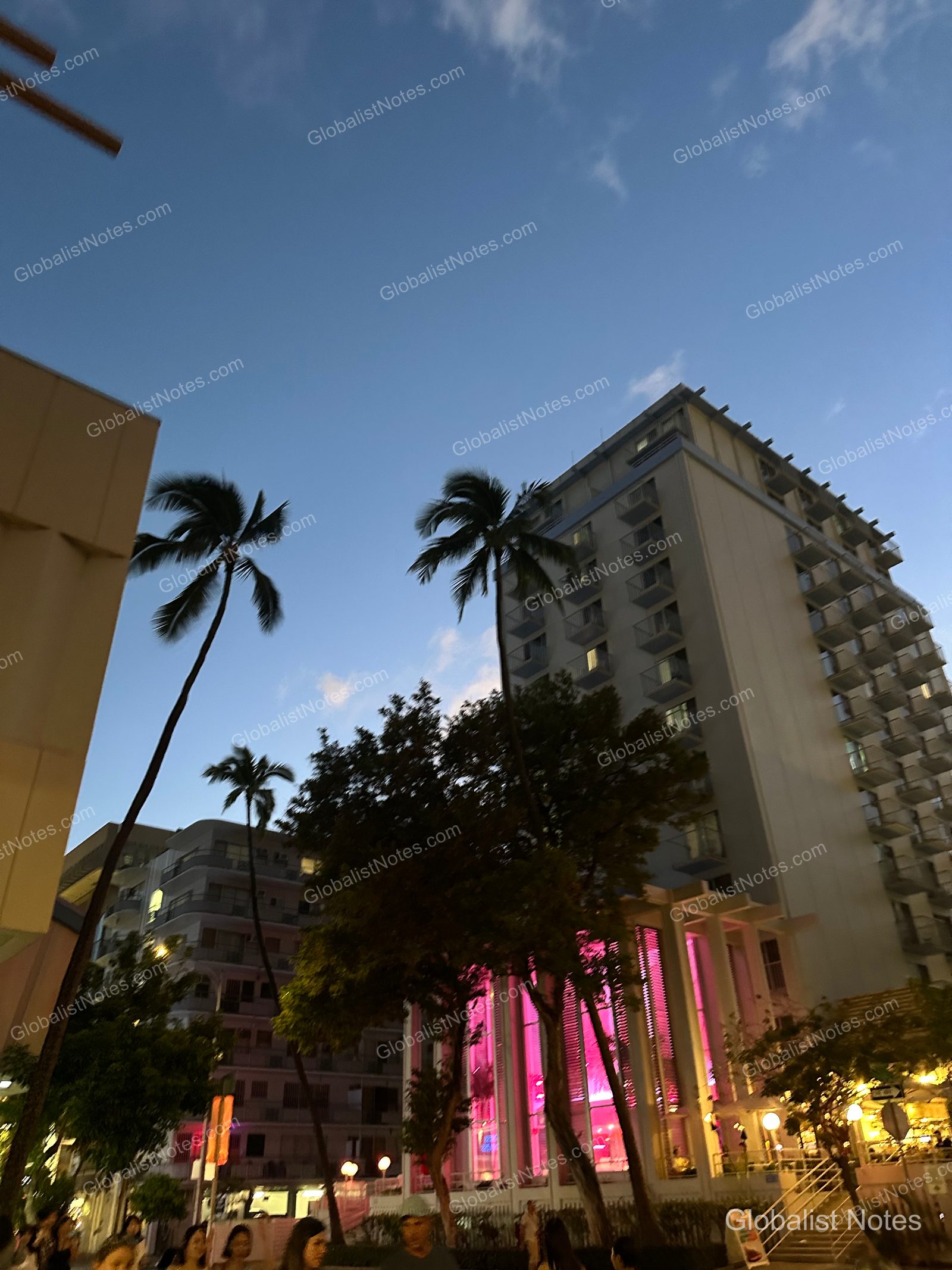 Exterior view of Hyatt Centric Waikiki Beach in Honolulu, Hawaii. The hotel building and palm trees are silhouetted against a blue twilight sky, while the ground-floor lobby is illuminated with a bright pink glow. Pedestrians are visible on the street in front of the building.