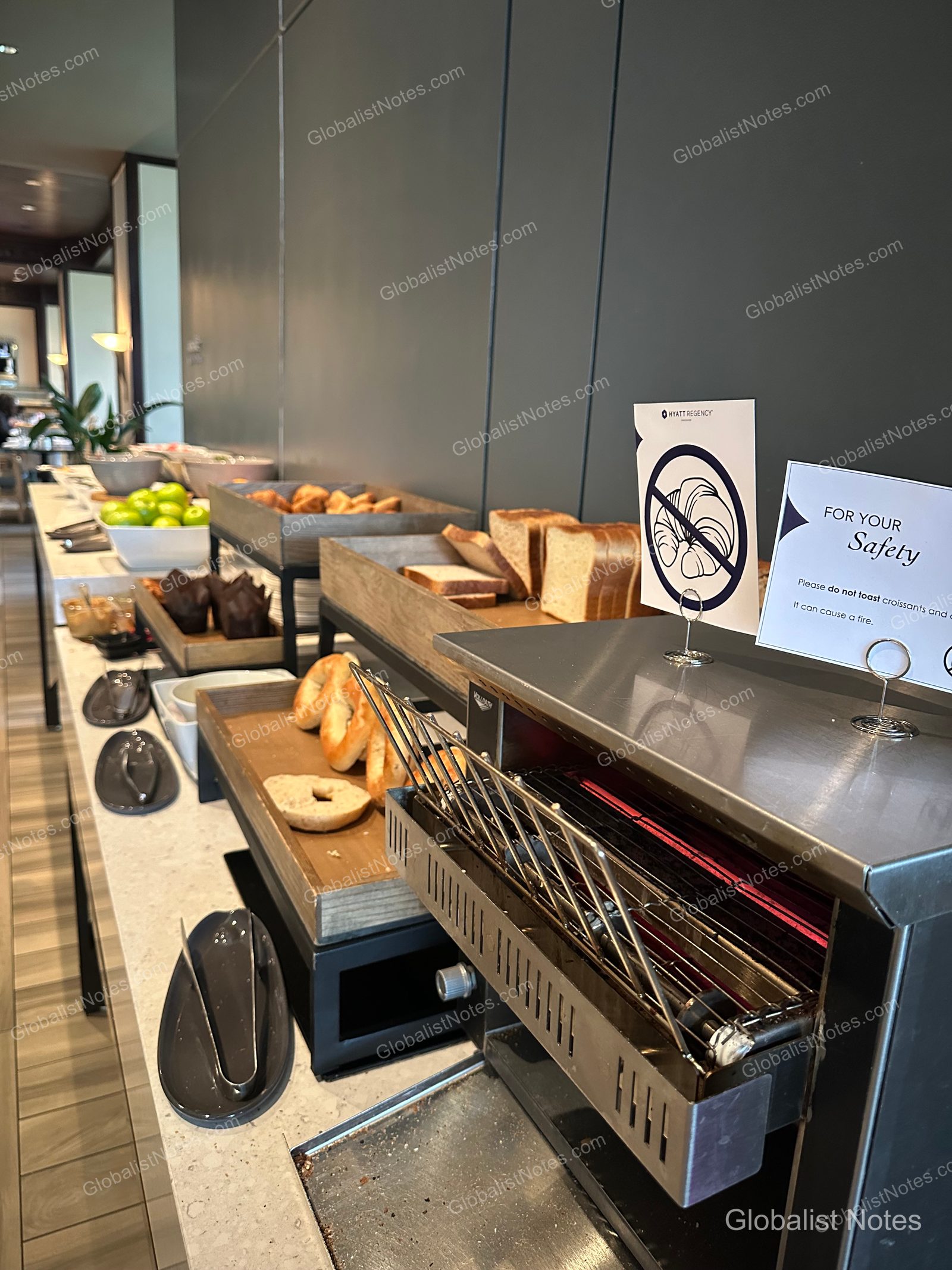 Bakery station at the Hyatt Regency Vancouver breakfast buffet. A safety sign on the toaster warns guests not to toast croissants, while the wooden trays behind feature an assortment of sliced bread, bagels, and muffins.