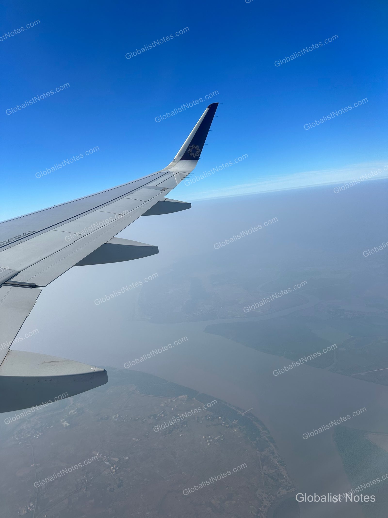 An aerial view of Mumbai from an airplane window, showing the coastline and river systems under a clear blue sky, with the wingtip visible in the upper left.