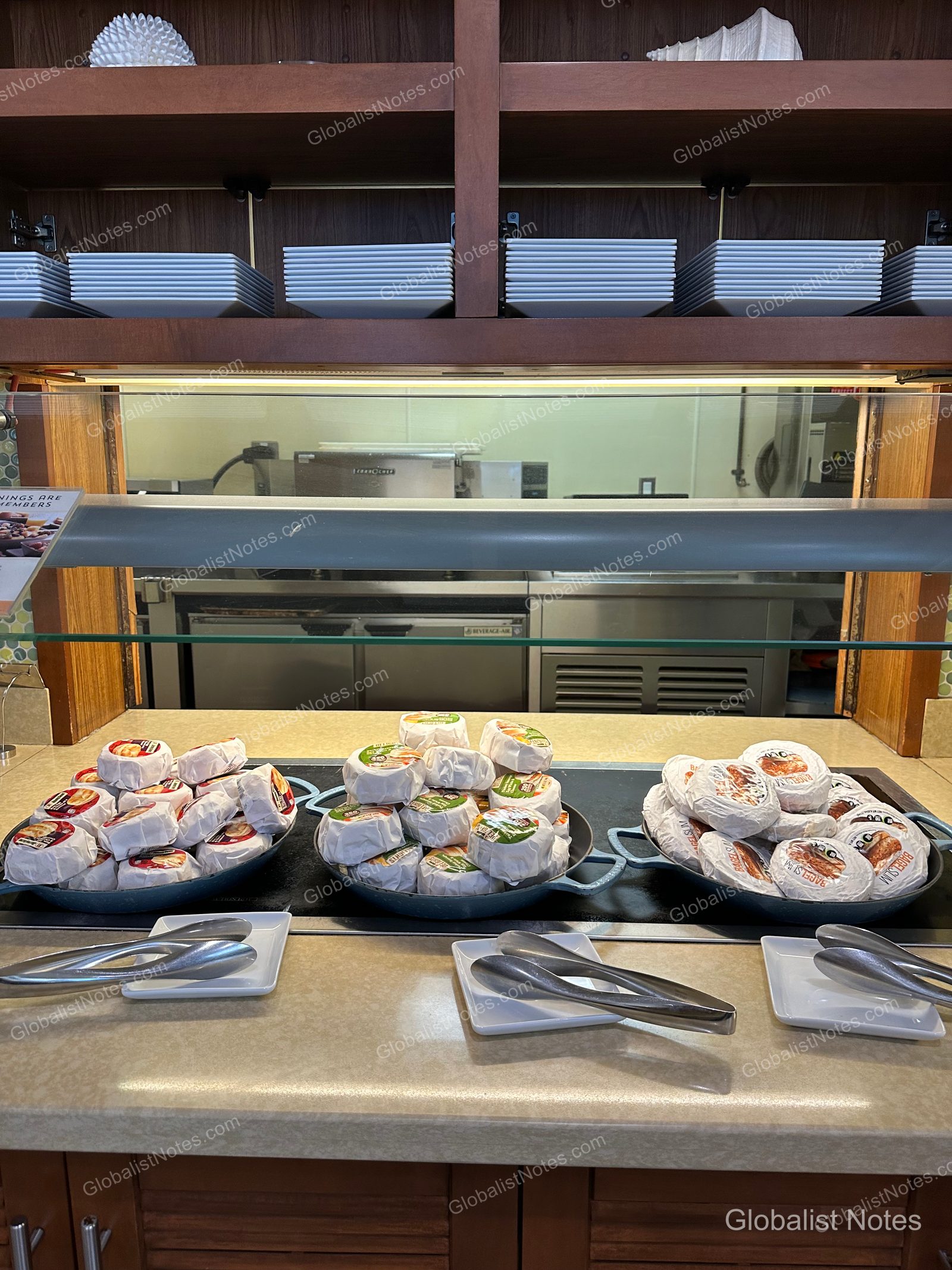 Individually wrapped bagels displayed on three round plates at the Hyatt Place Waikiki Beach breakfast buffet, with stacks of white plates on the shelf above.