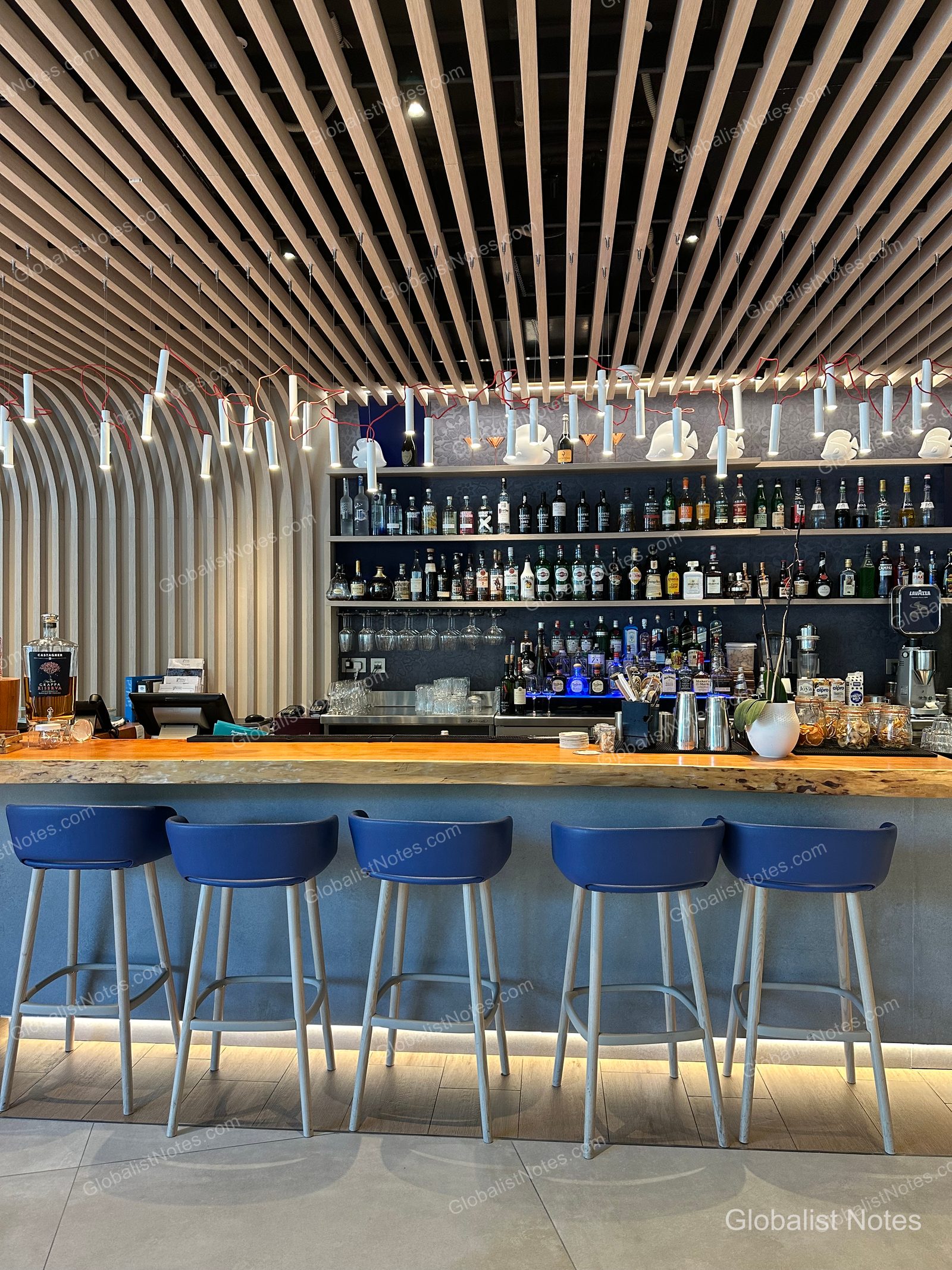 The bar at Hyatt Regency Malta, featuring curved wooden ceilings and walls, a long wooden counter, and five blue bar stools. The back bar is stocked with spirits, and elongated light fixtures hang from the ceiling at irregular intervals.