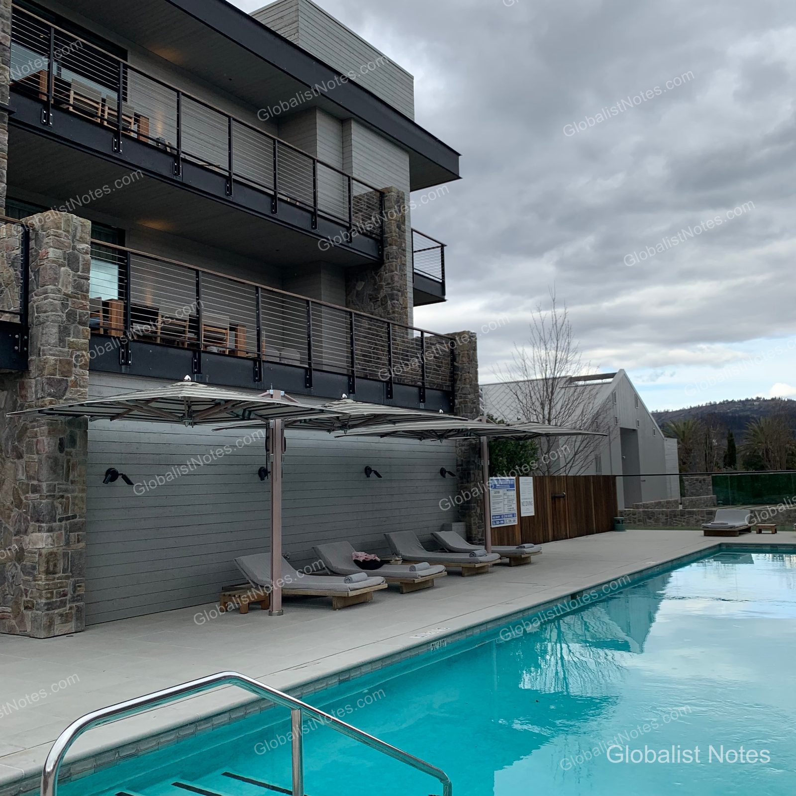 Outdoor pool view at Alila Napa Valley in St. Helena, USA. Sun loungers and umbrellas are arranged on the pool deck, with the balconies of the 2 Queen Beds Vineyard View Balcony rooms visible in the background.