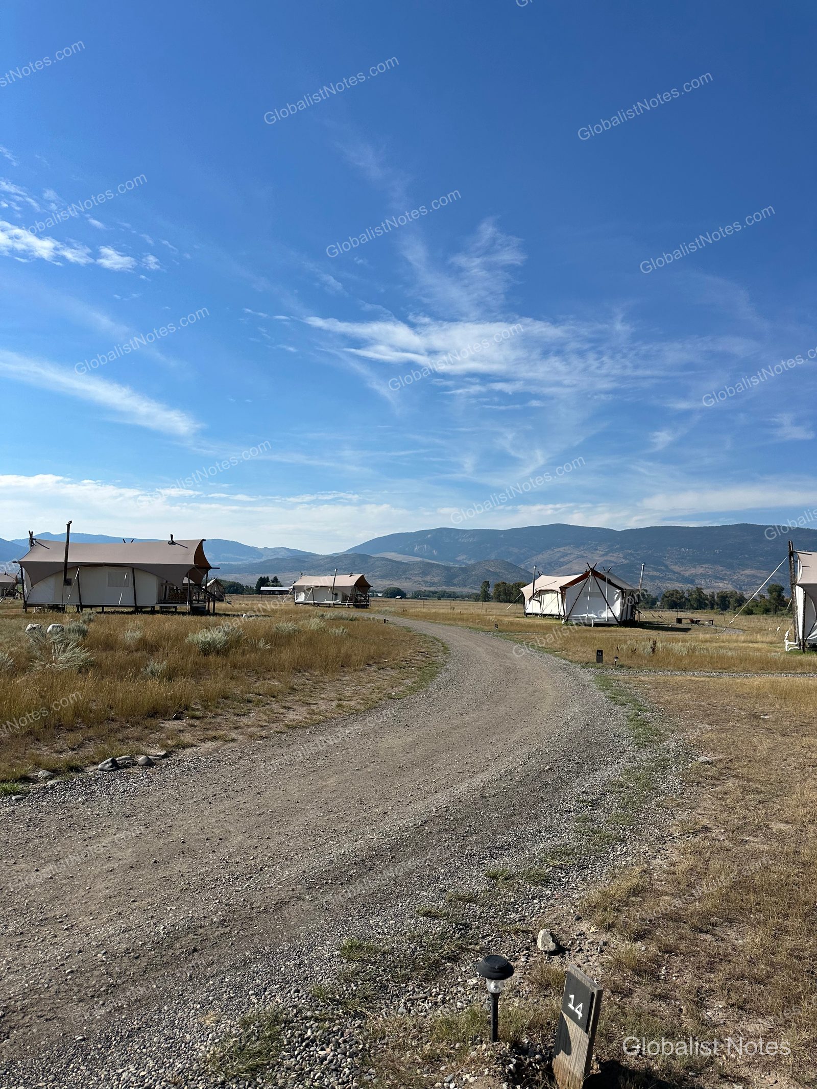 米国パインクリークのUnder Canvas North Yellowstone。広大な草原にテントが並び、背景には山脈が広がっています。手前の砂利道には14番テントを示す看板が見えます。