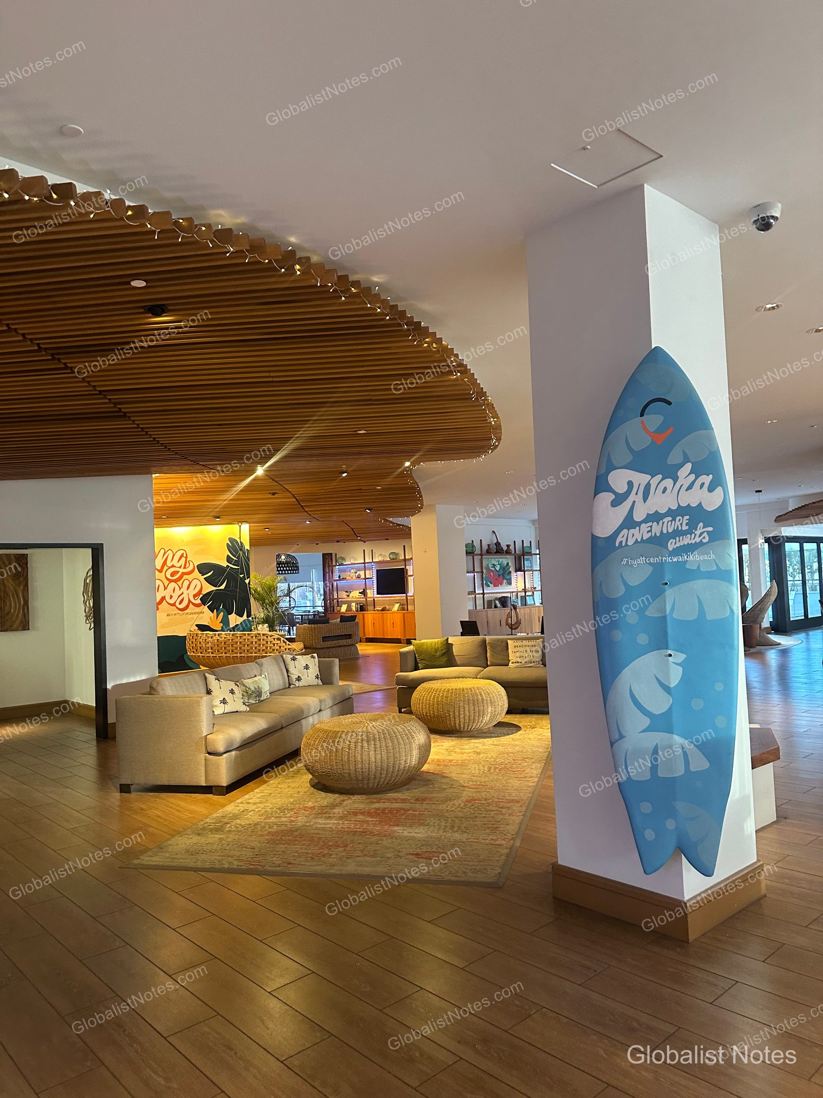 Lobby view at Hyatt Centric Waikiki Beach. The space features white pillars adorned with blue surfboard accents, a ceiling finished with wooden slats, and a seating area with sofas and round rattan stools. The floor is finished with wood-patterned tile, with additional wall decor and furniture placed toward the back.
