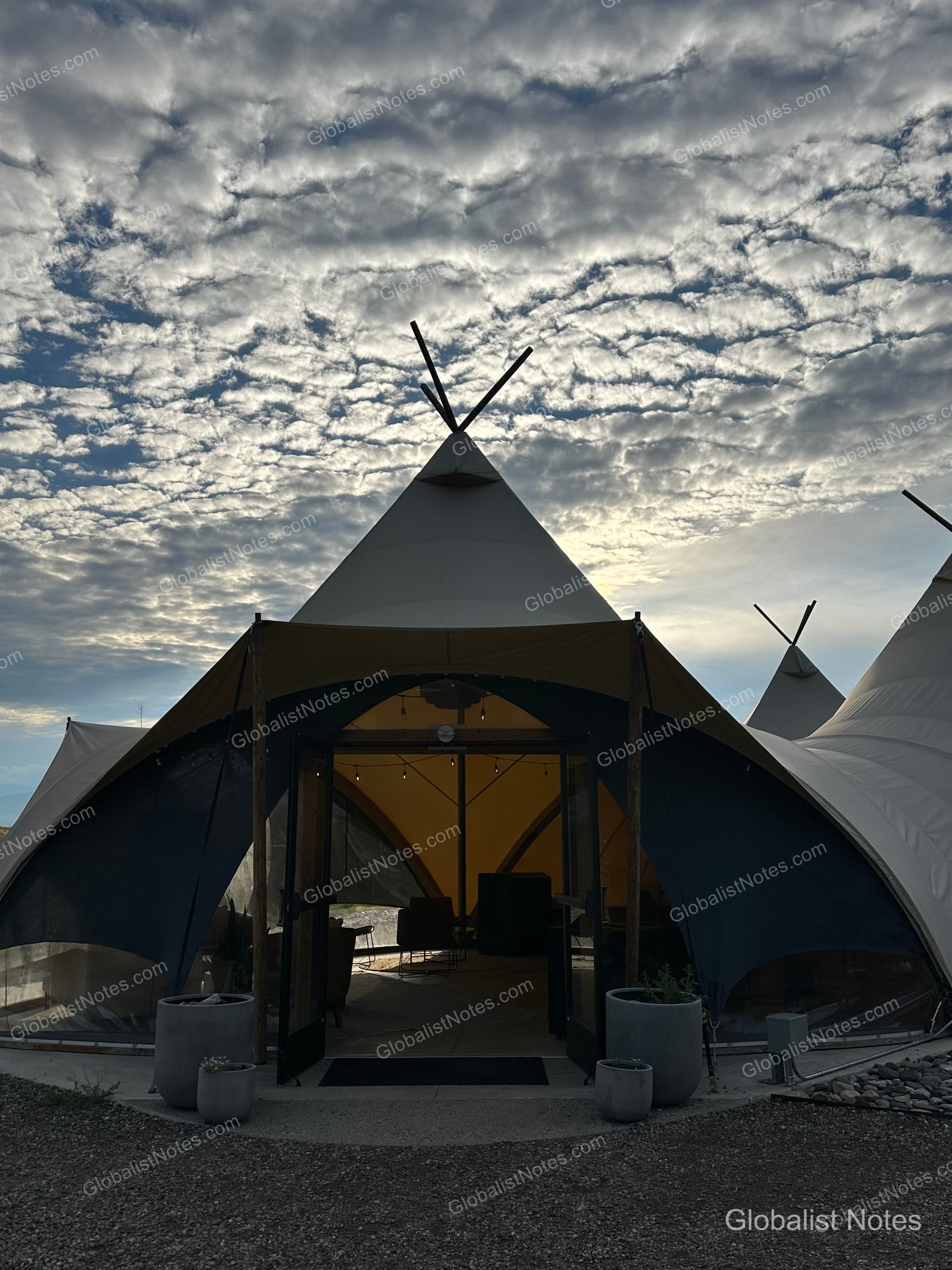 Under Canvas North Yellowstoneのテント宿泊施設の全景。とんがり屋根のテントの入り口が開き、内部に明かりが灯っている。背景には雲に覆われた空が広がる。