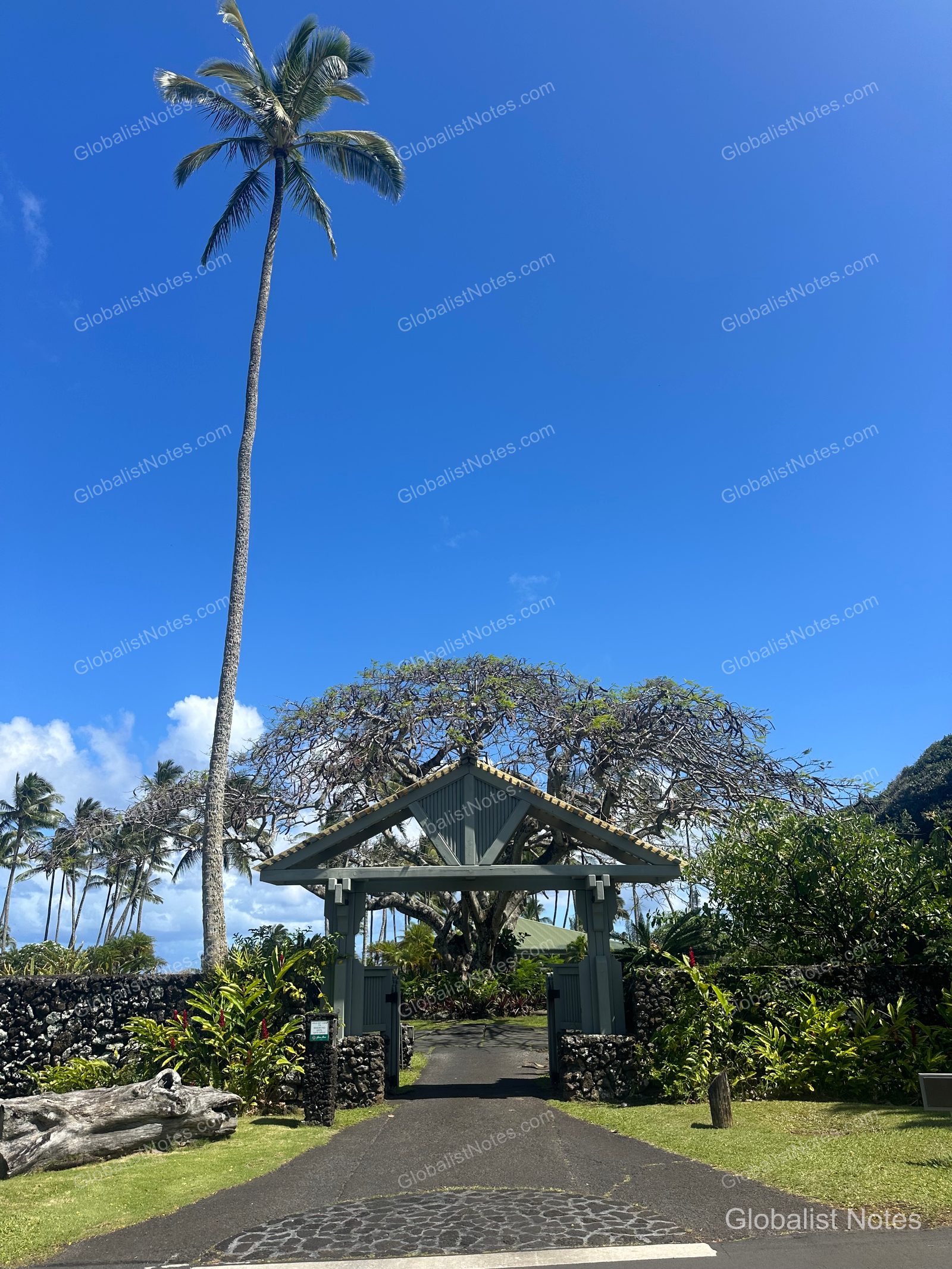 The entrance to Hana-Maui Resort on Maui, Hawaii. A traditional wooden resort gate stands at the end of the road, framed by tall palm trees, stone walls, and lush greenery under a clear blue sky.