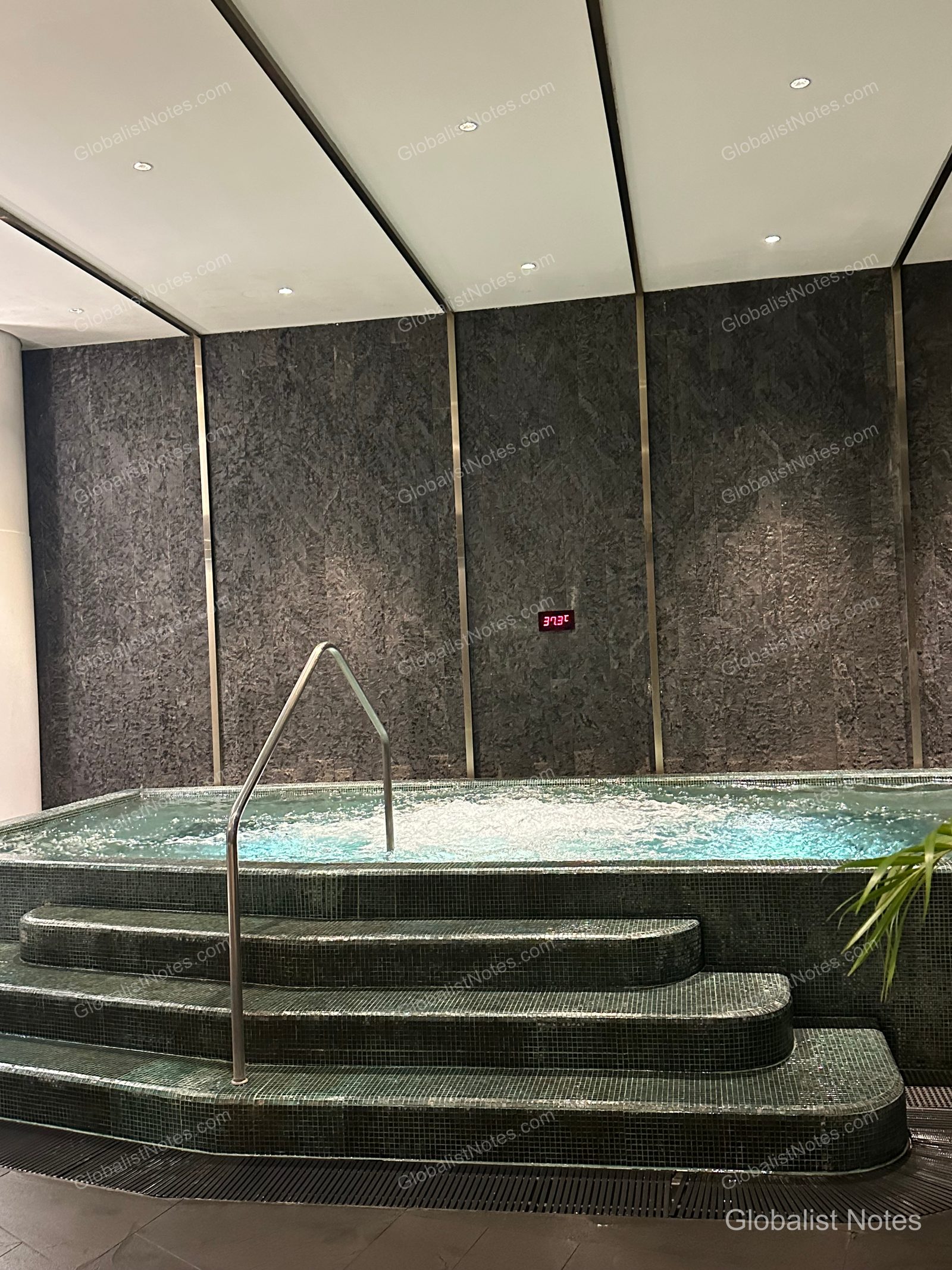 Heated jacuzzi inside the Grand Hyatt Jeju indoor pool. The jacuzzi features a tiered design against a dark stone wall, with bubbles rising to the surface and a digital display on the wall showing a temperature of 37.3°C.