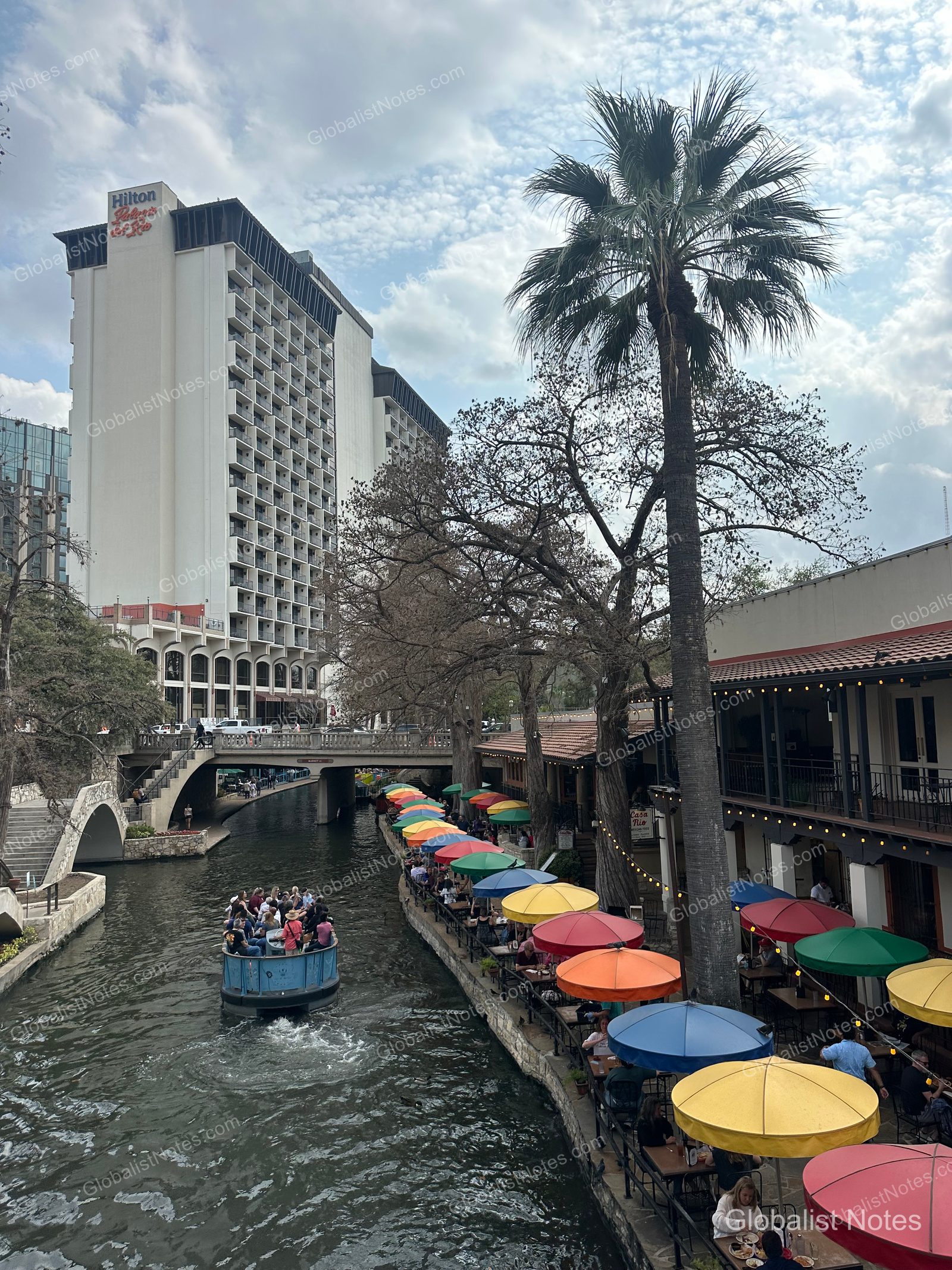 The San Antonio River Walk canal featuring outdoor dining areas with colorful umbrellas and a passing tour boat. The hotel building is visible on the left, with small bridges crossing the canal and palm trees lining the urban walkway.