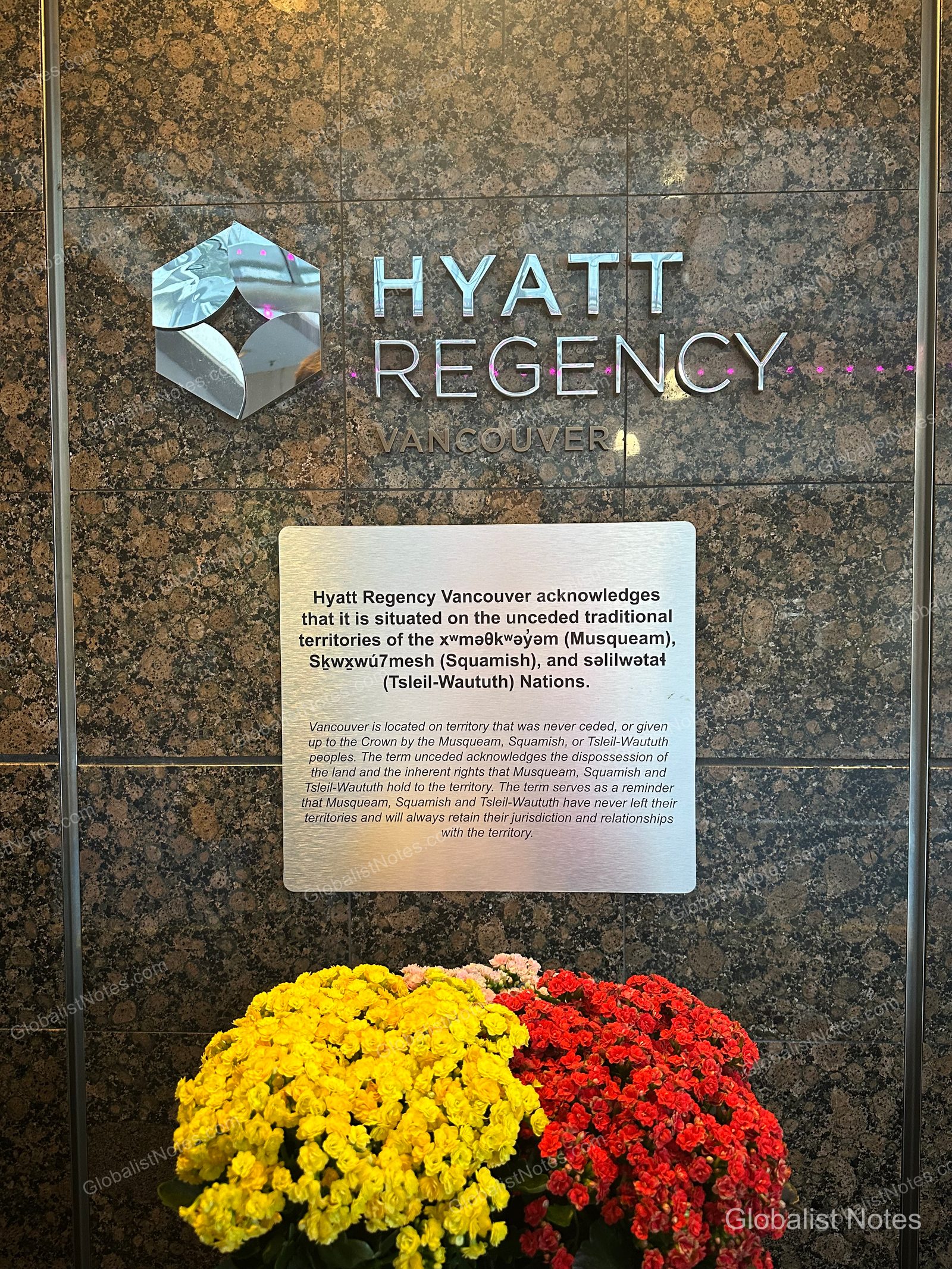 The Hyatt Regency Vancouver logo mounted on a brown marble wall in the lobby, alongside a plaque acknowledging the traditional territory of the Musqueam, Squamish, and Tsleil-Waututh Nations. Yellow and red floral arrangements are placed below.