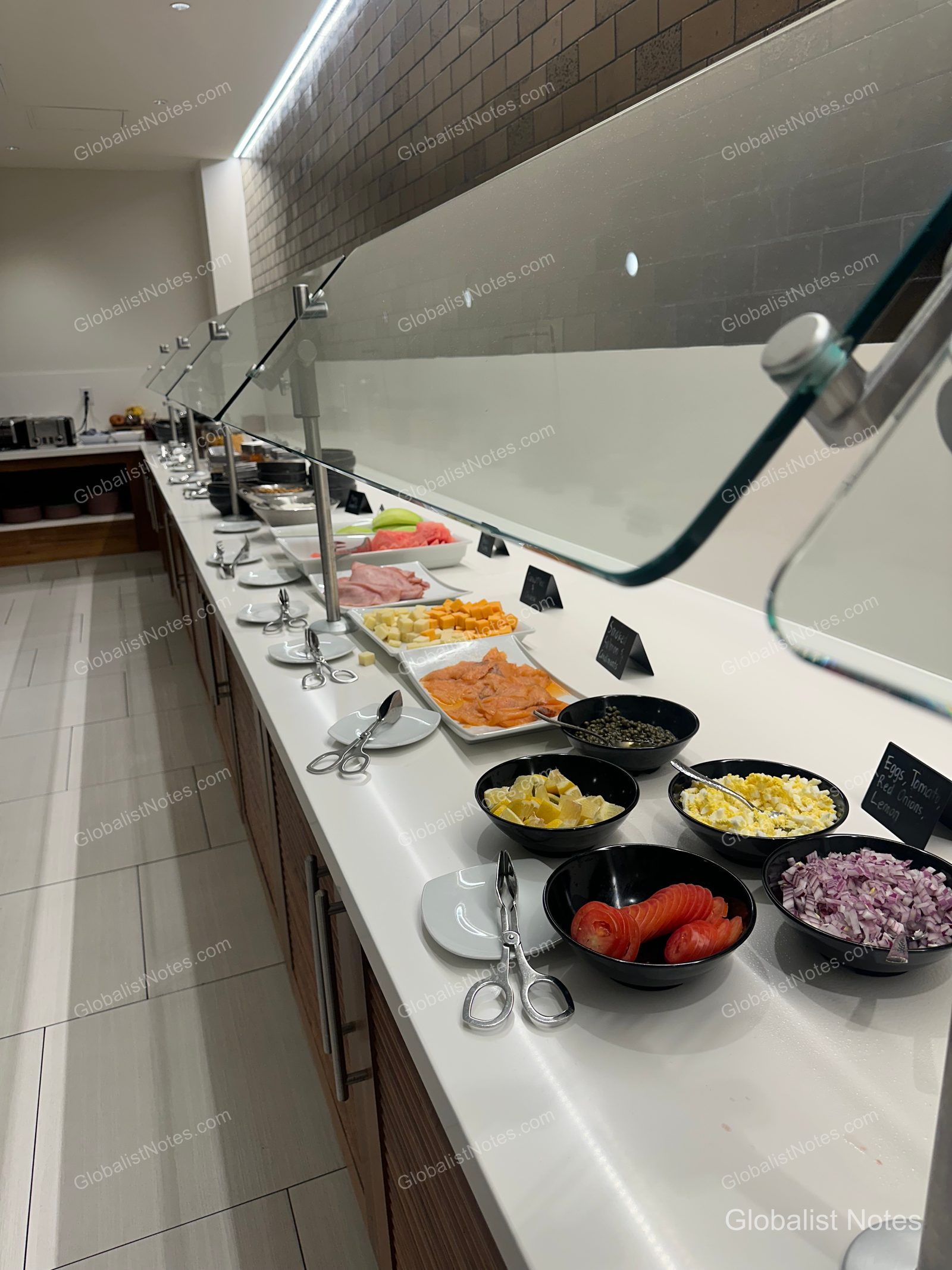 Cold food section at the Hyatt Regency Los Angeles International Airport breakfast buffet. Smoked salmon, cheeses, cubed fruit, sliced tomatoes, and diced red onions are neatly displayed in black bowls and square platters under a glass sneeze guard on a long white table.