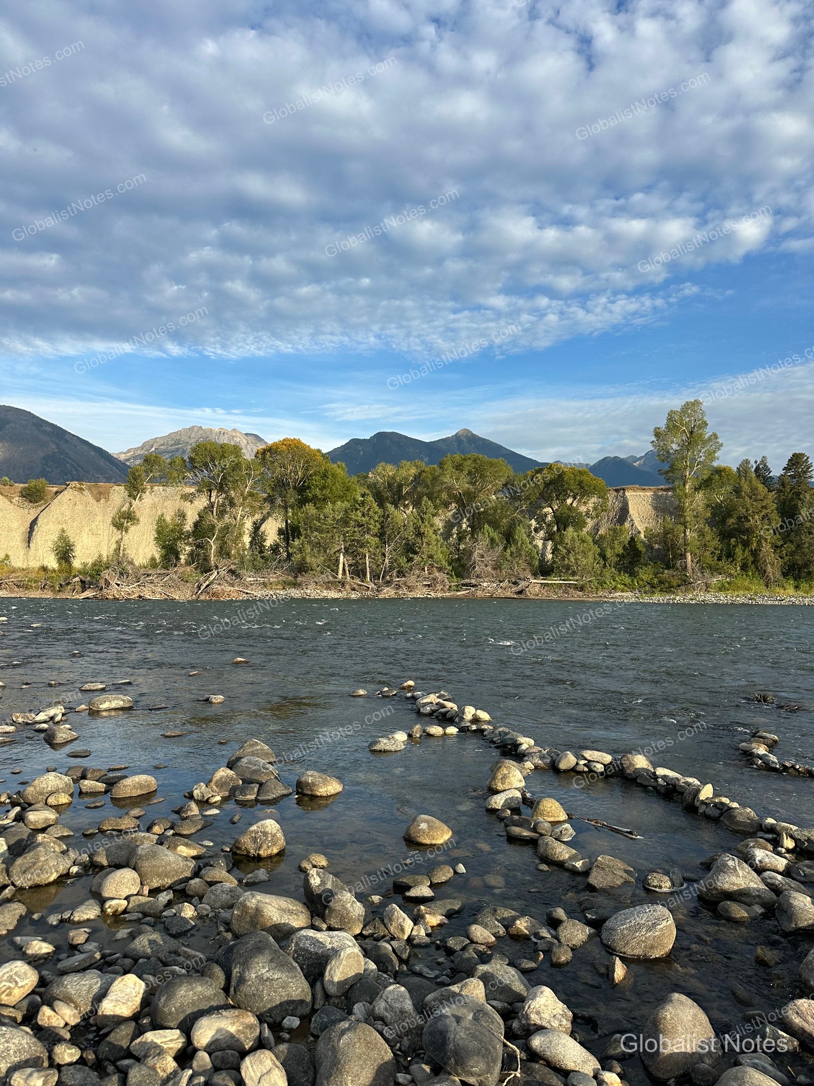 Under Canvas North Yellowstone近くを流れる川の風景。晴天の下、山々と森を背景に、浅い川底の石の間を水が流れています。
