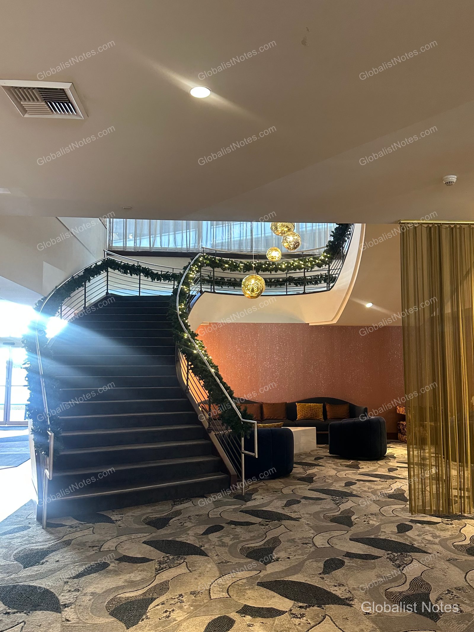 Curved staircase in the lobby of the Hyatt Regency Los Angeles International Airport. The railing is decorated with greenery, spherical light fixtures hang from the ceiling, and a seating area with sofas and tables is visible to the side.