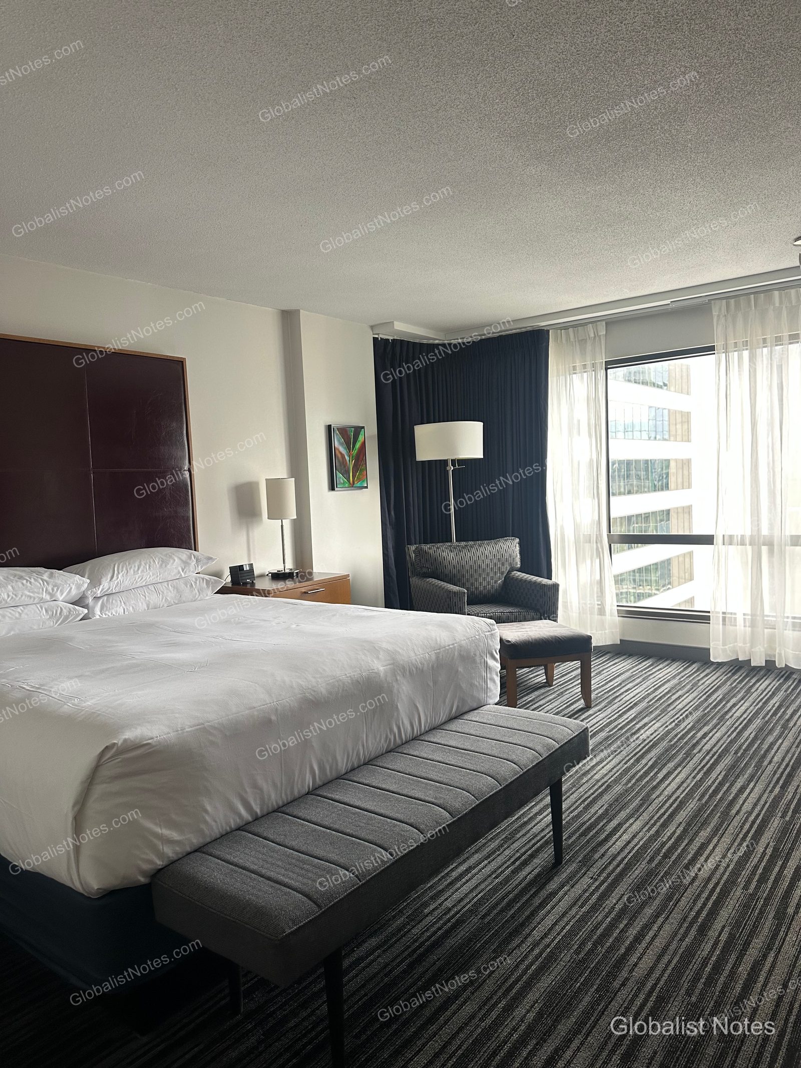Interior of a Deluxe Suite at Hyatt Regency Vancouver. A gray bench is placed at the foot of the king bed, with an armchair, ottoman, and floor lamp arranged by the window. The room features striped carpeting and views of surrounding downtown buildings.