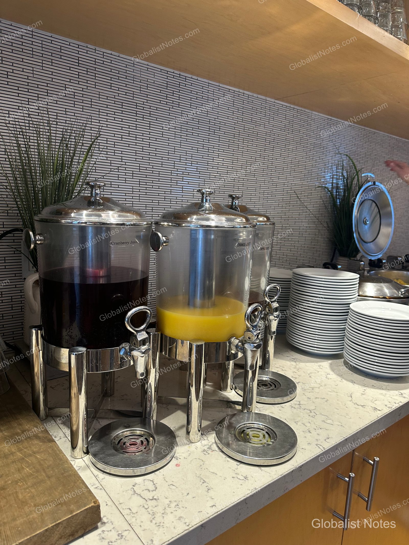 Manchester Grand Hyatt San Diego 自助早餐區的三台不鏽鋼飲料機，分別供應蔓越莓汁、柳橙汁與蘋果汁，一旁整齊堆疊著白色餐盤。