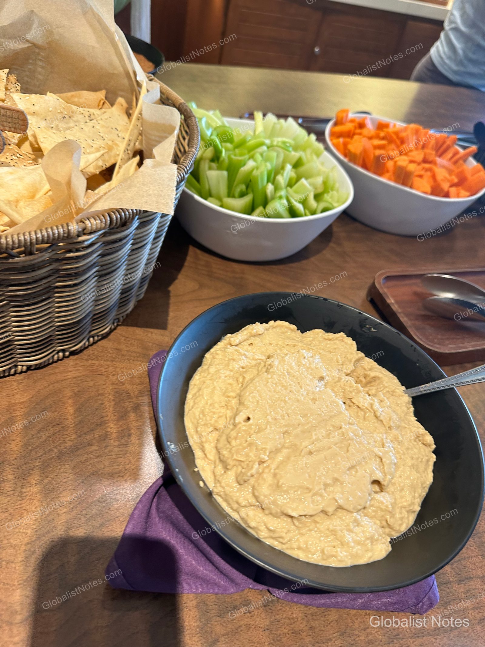 Snacks served in the lounge at Hyatt Regency Maui Resort and Spa. A basket of nacho chips is on the left, a white bowl containing celery and carrot sticks is on the right, and a black bowl of hummus is in the foreground.
