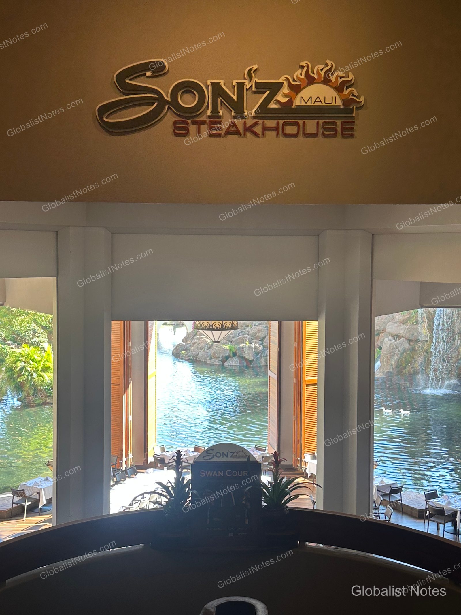 Entrance to Son'z Steakhouse at Hyatt Regency Maui Resort and Spa. The restaurant logo is displayed above the entrance, and the open windows look out onto an outdoor pond, waterfall, and terrace dining area.