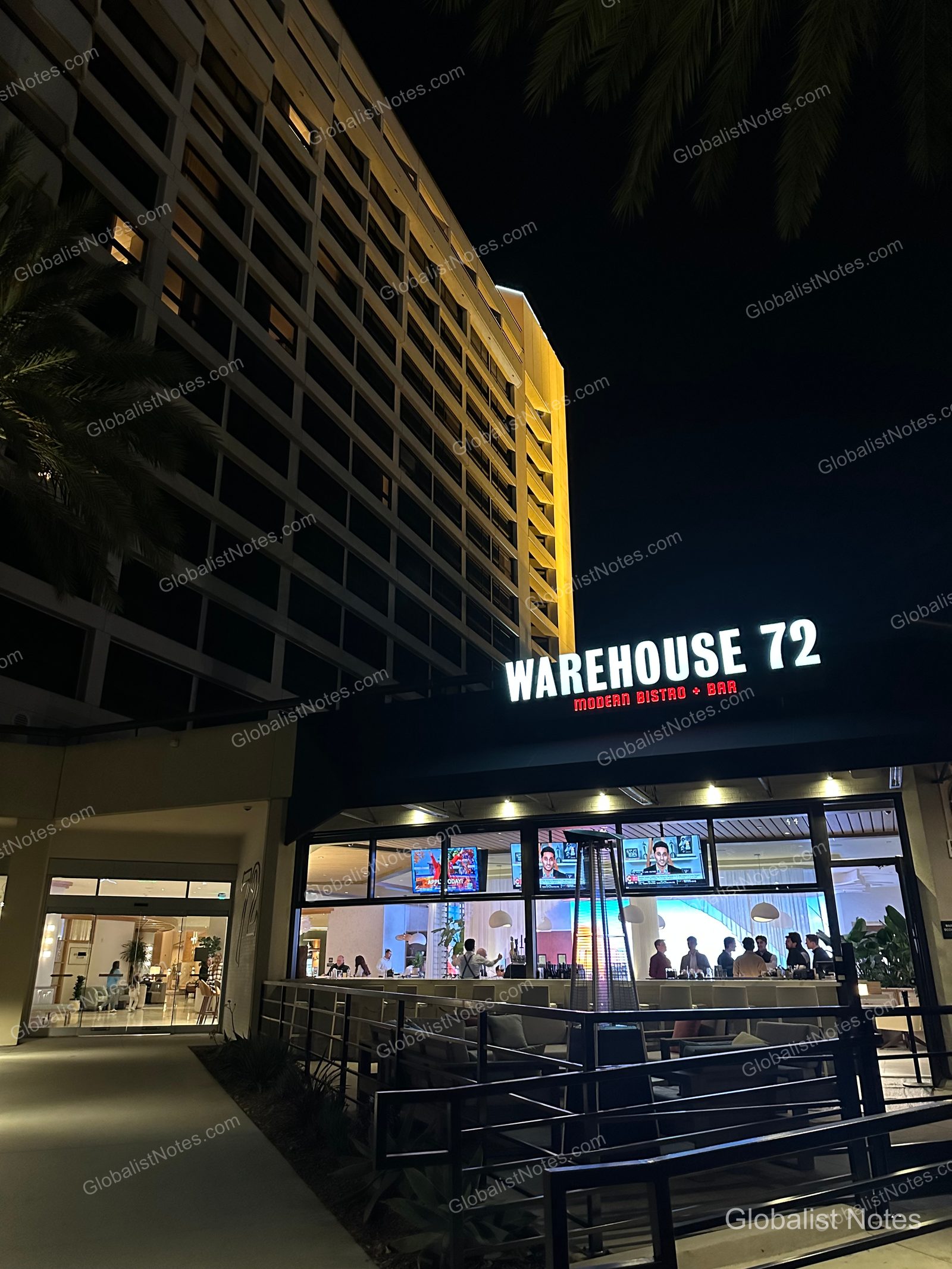 夜間點燈的 Hyatt Regency Irvine 建築旁，可見 Warehouse 72 餐廳招牌與戶外露台座位。透過落地窗可見室內吧台與電視，建築外牆在暖黃燈光下顯得格外明亮。