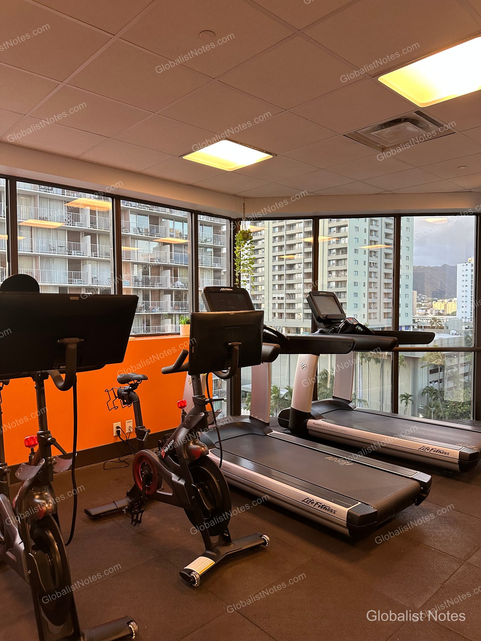Inside the fitness center at Hyatt Centric Waikiki Beach. Peloton stationary bikes and Life Fitness treadmills are arranged in front of floor-to-ceiling windows, which offer views of downtown Honolulu buildings and the mountains.