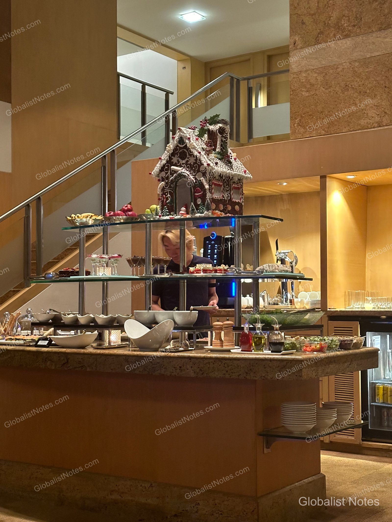 A large gingerbread house centerpiece in the Grand Hyatt Mumbai lounge, with a buffet of desserts and salads displayed below.