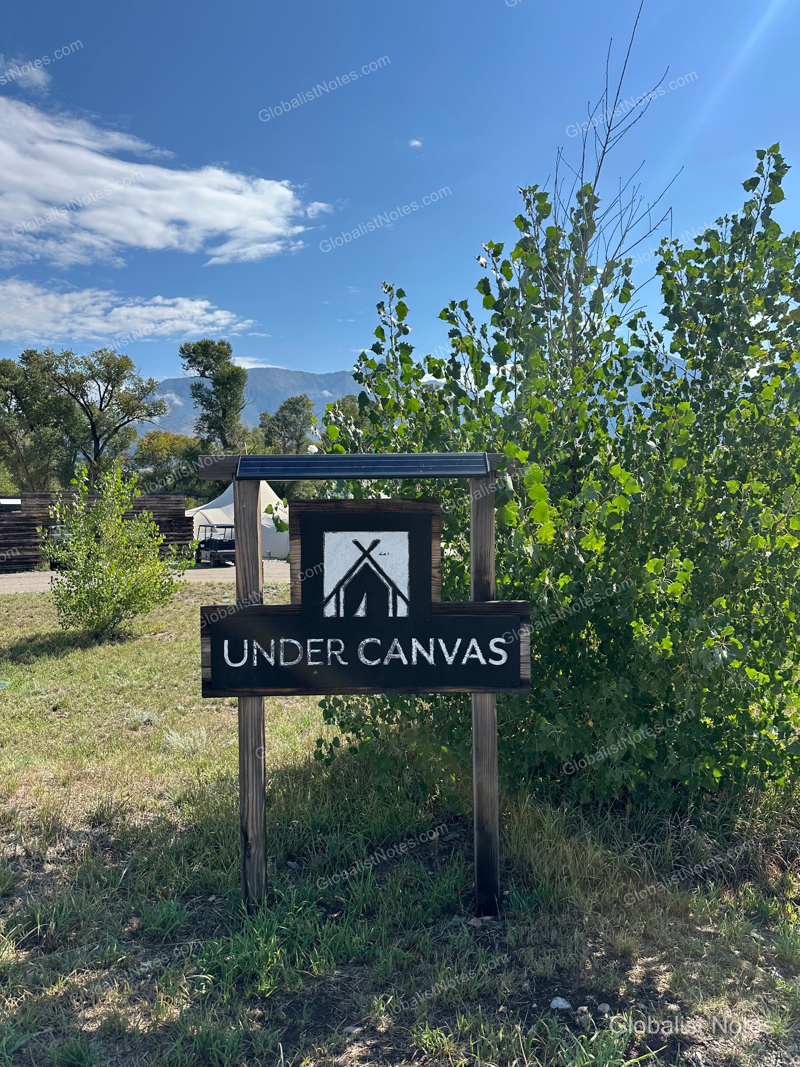パインクリークのUNDER CANVAS North Yellowstone入り口にある木製の看板。黒地に白いテントのロゴと「UNDER CANVAS」の文字が刻まれ、背景には青空と山、テント施設が見えます。