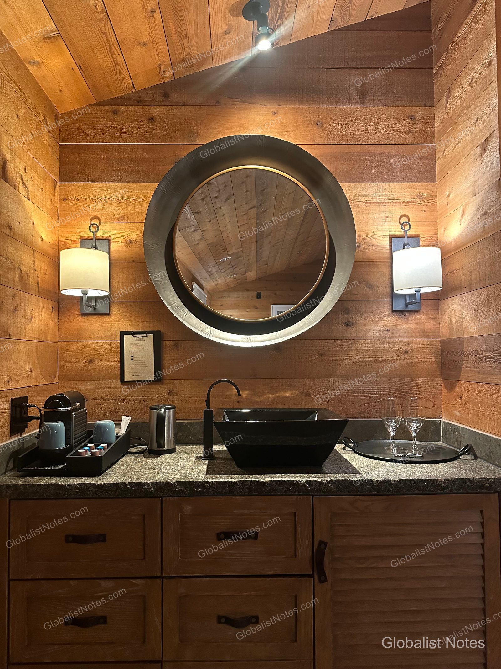 Bathroom in an Alila Ventana Big Sur Junior Suite featuring a round mirror and a black square sink. The walls are finished with warm wood paneling, flanked by lighting, with a Nespresso machine and glassware neatly arranged next to the sink.