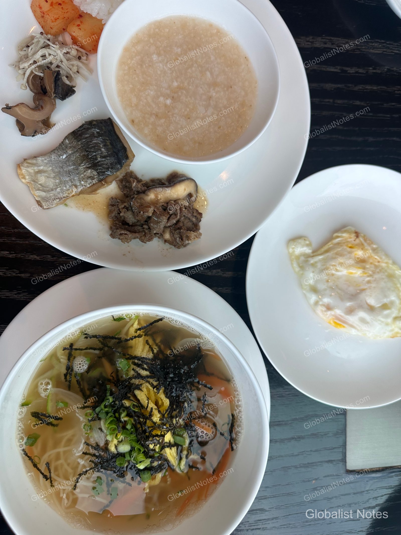 Korean breakfast options at the Grand Hyatt Jeju buffet. A large white plate holds porridge, grilled fish, bulgogi, cubed radish kimchi, and seasoned vegetables, accompanied by a side plate of fried eggs and a bowl of warm noodles topped with garnishes and seaweed.