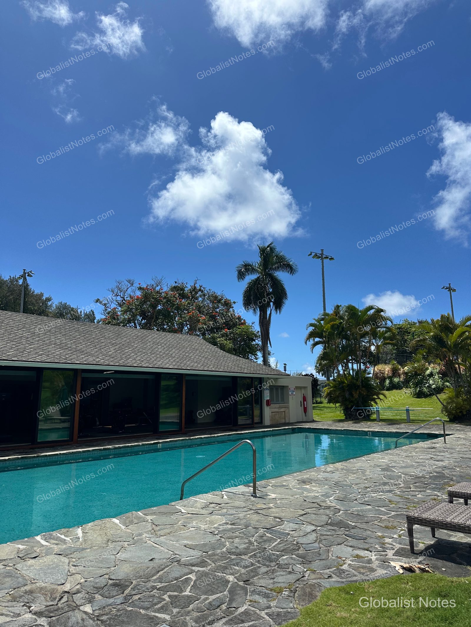 View of the outdoor pool at Hana-Maui Resort. The pool features a stone-finished deck, blue water, and is surrounded by palm trees and resort buildings under a clear sky.